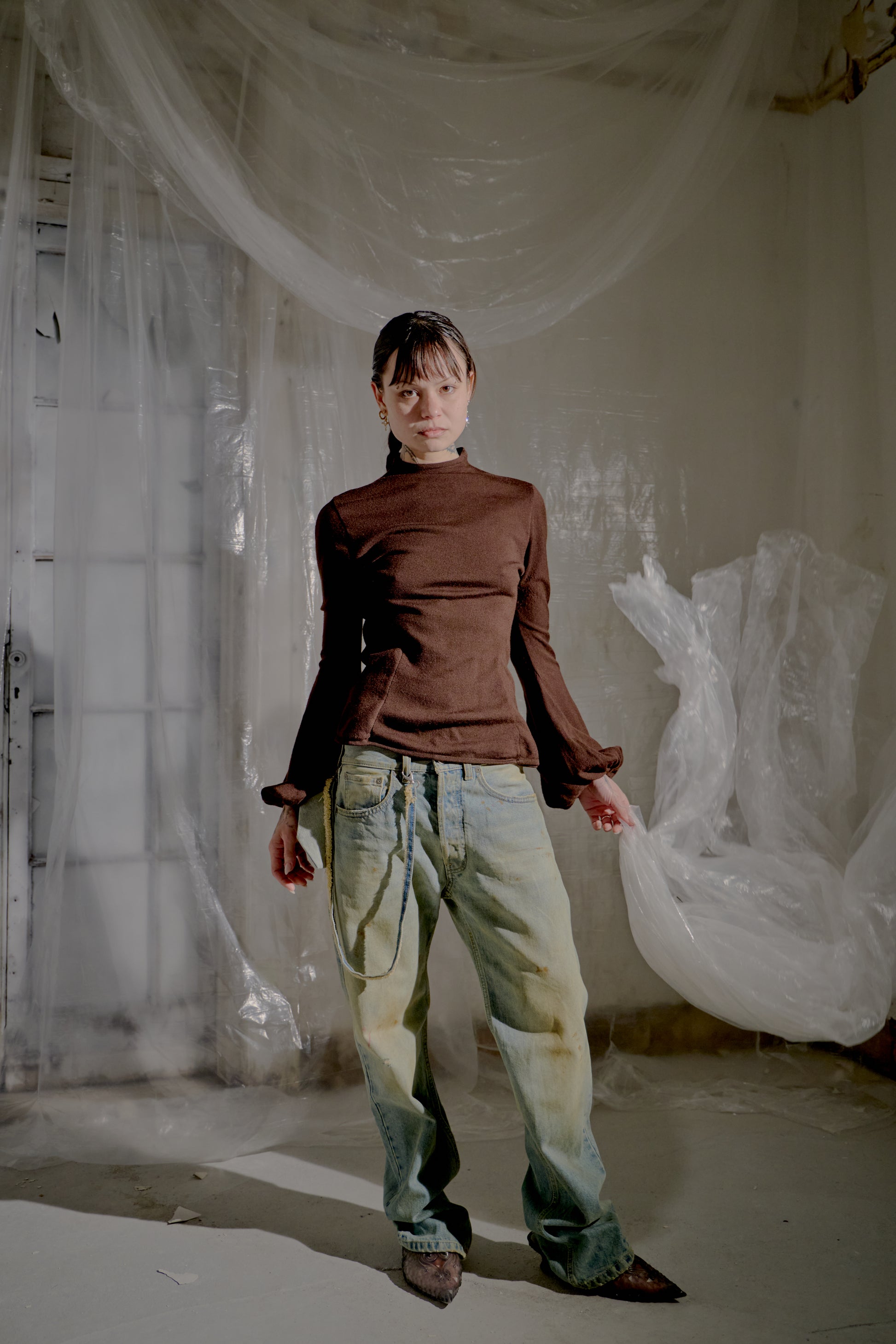 Person wearing a brown Alexander McQueen turtleneck and light-coloured pants in a room with white plastic sheeting.