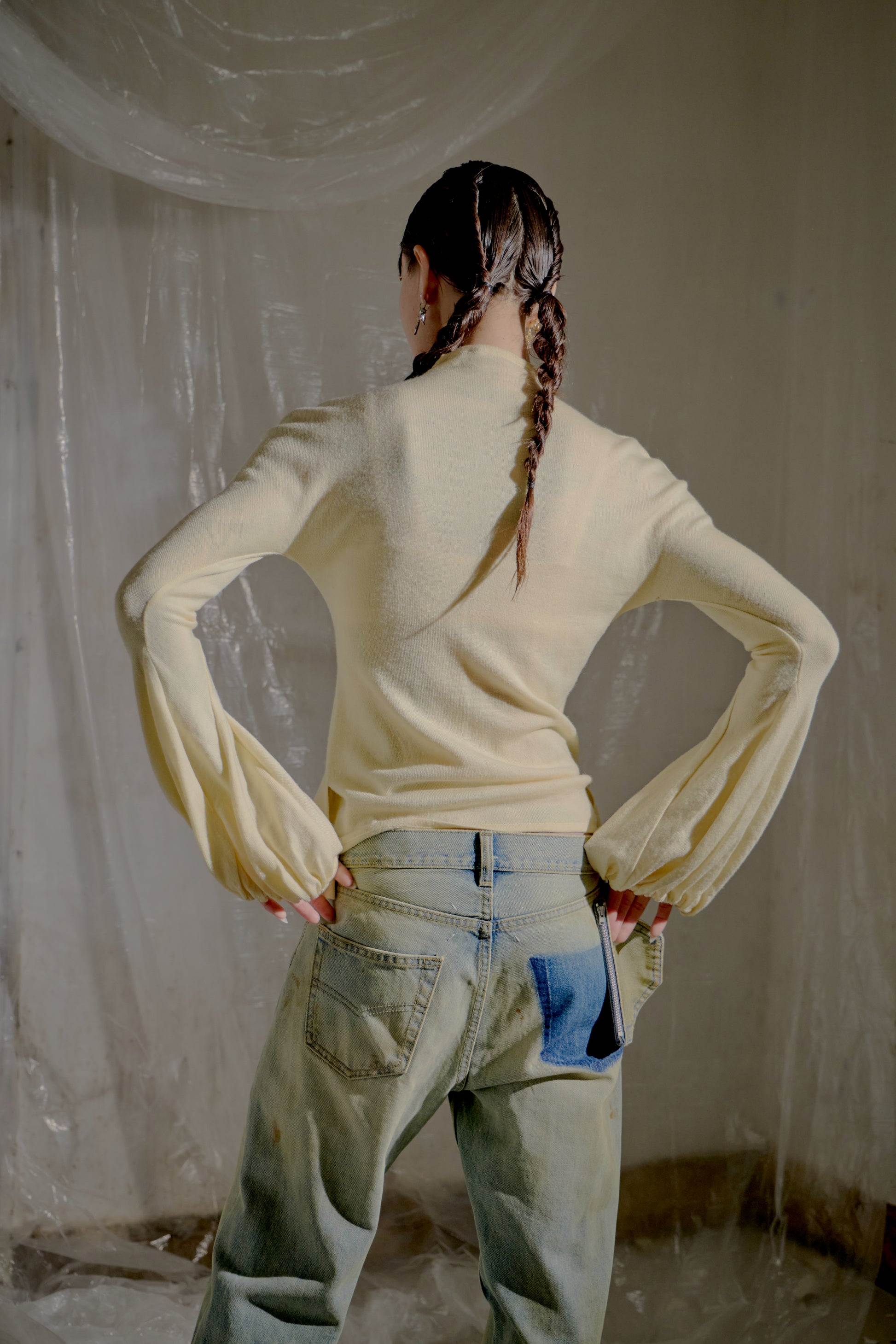 Person wearing a light-coloured Alexander McQueen sweater and jeans with a braid, standing in a room with white walls.
