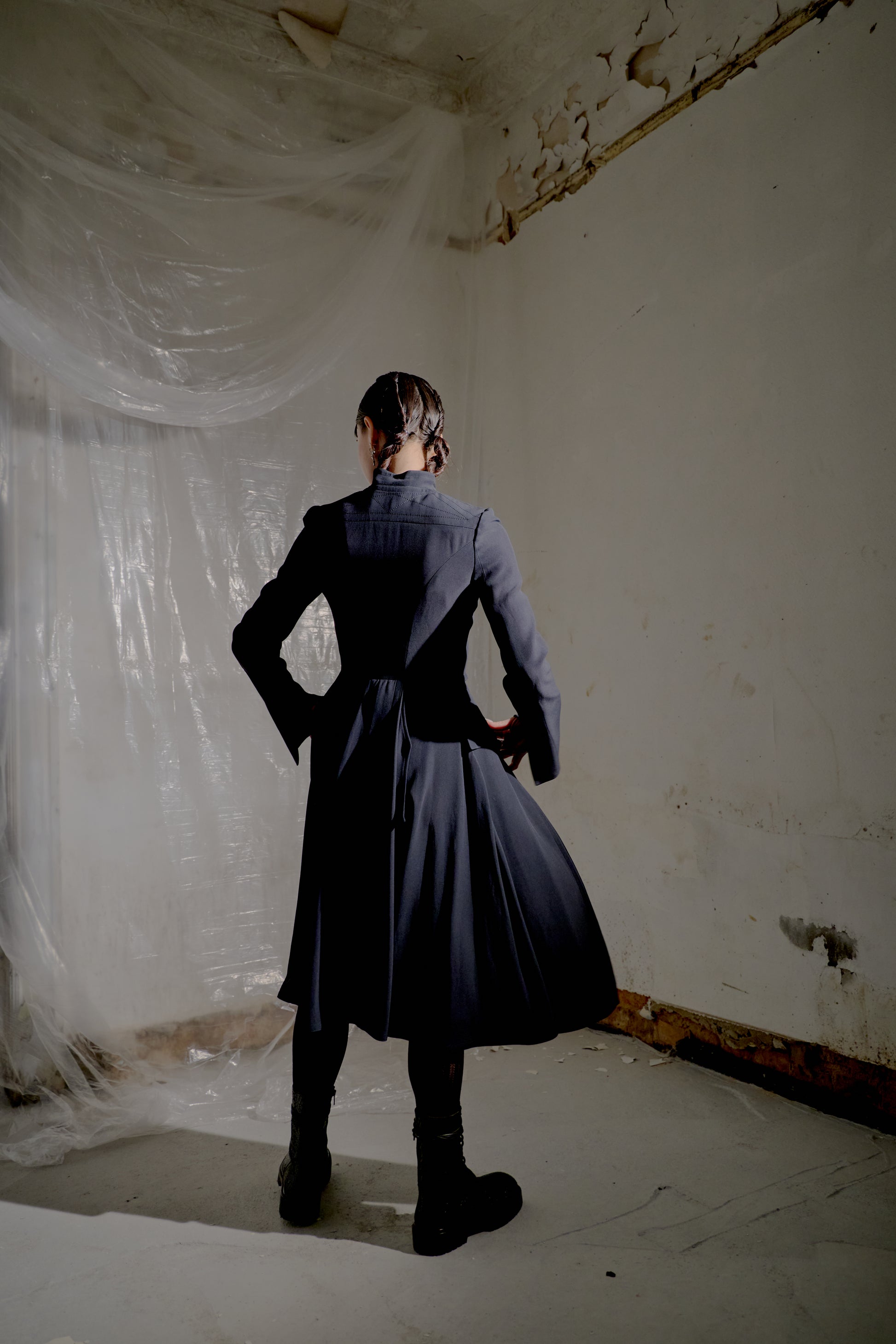 Person wearing a dark Alexander McQueen coat standing in a room with white plastic sheeting and peeling paint.