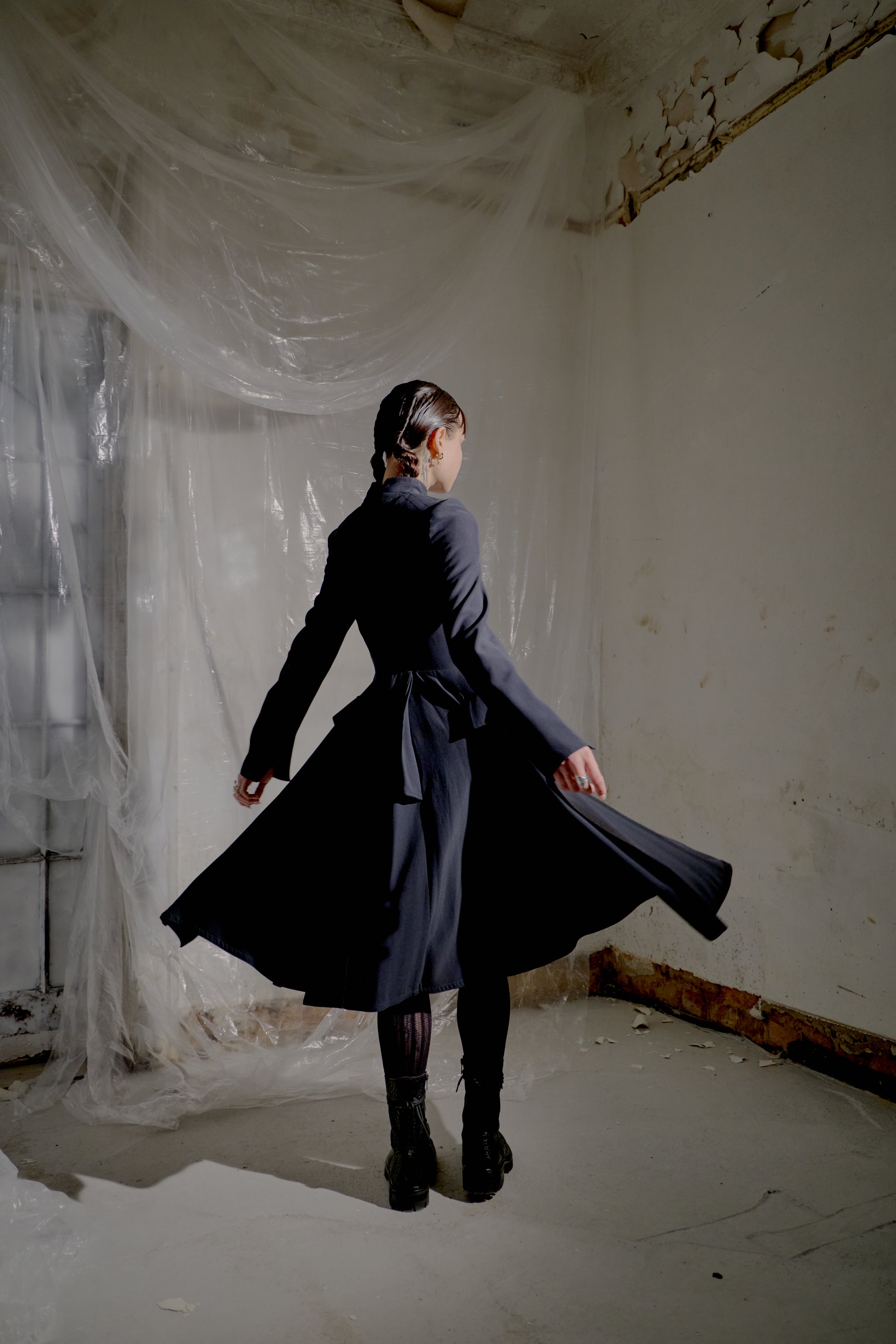 Person wearing a long dark Alexander McQueen coat in a room with white plastic sheeting on the walls.