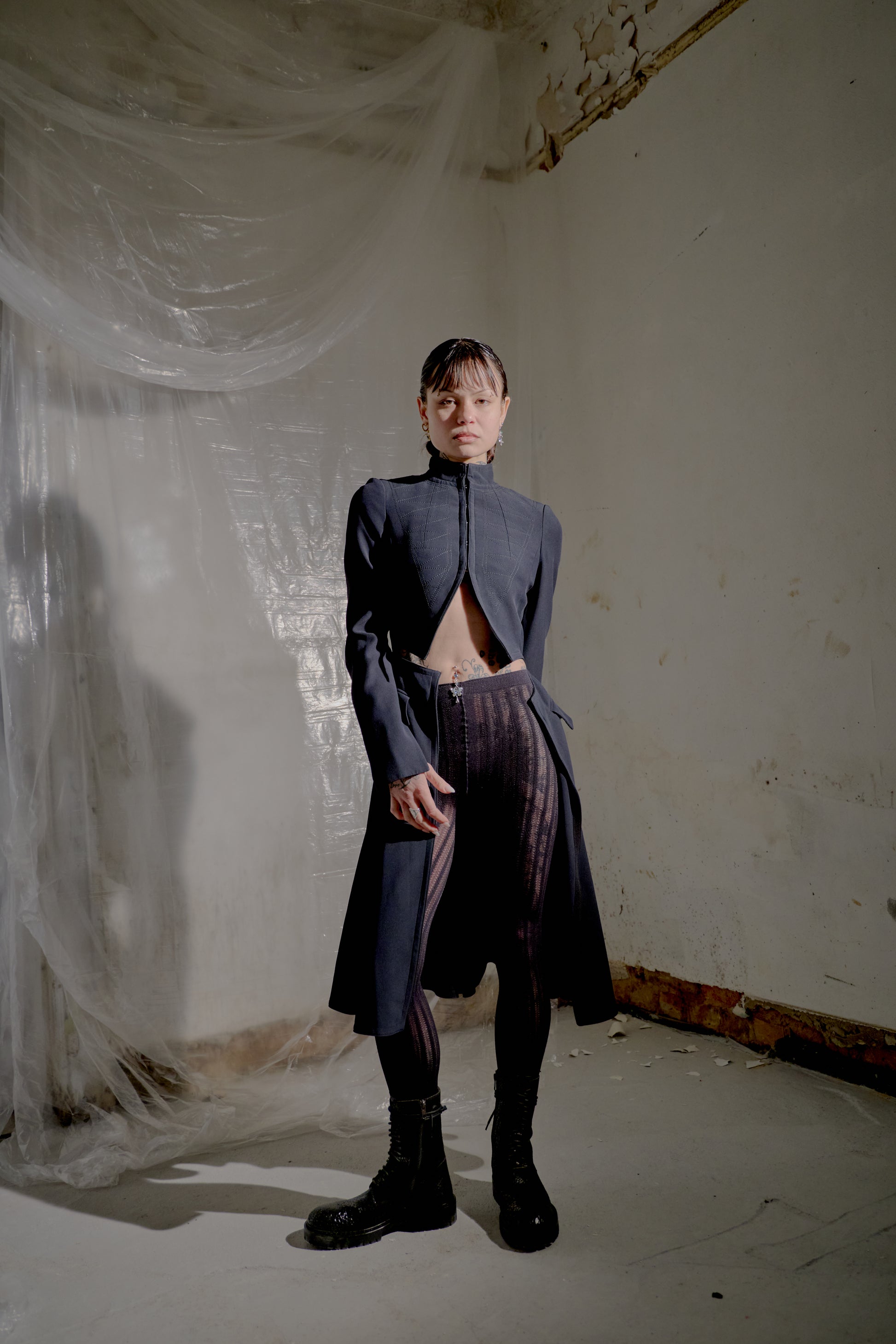 Person wearing a dark Alexander McQueen coat and pants in a room with white walls and a curtain.