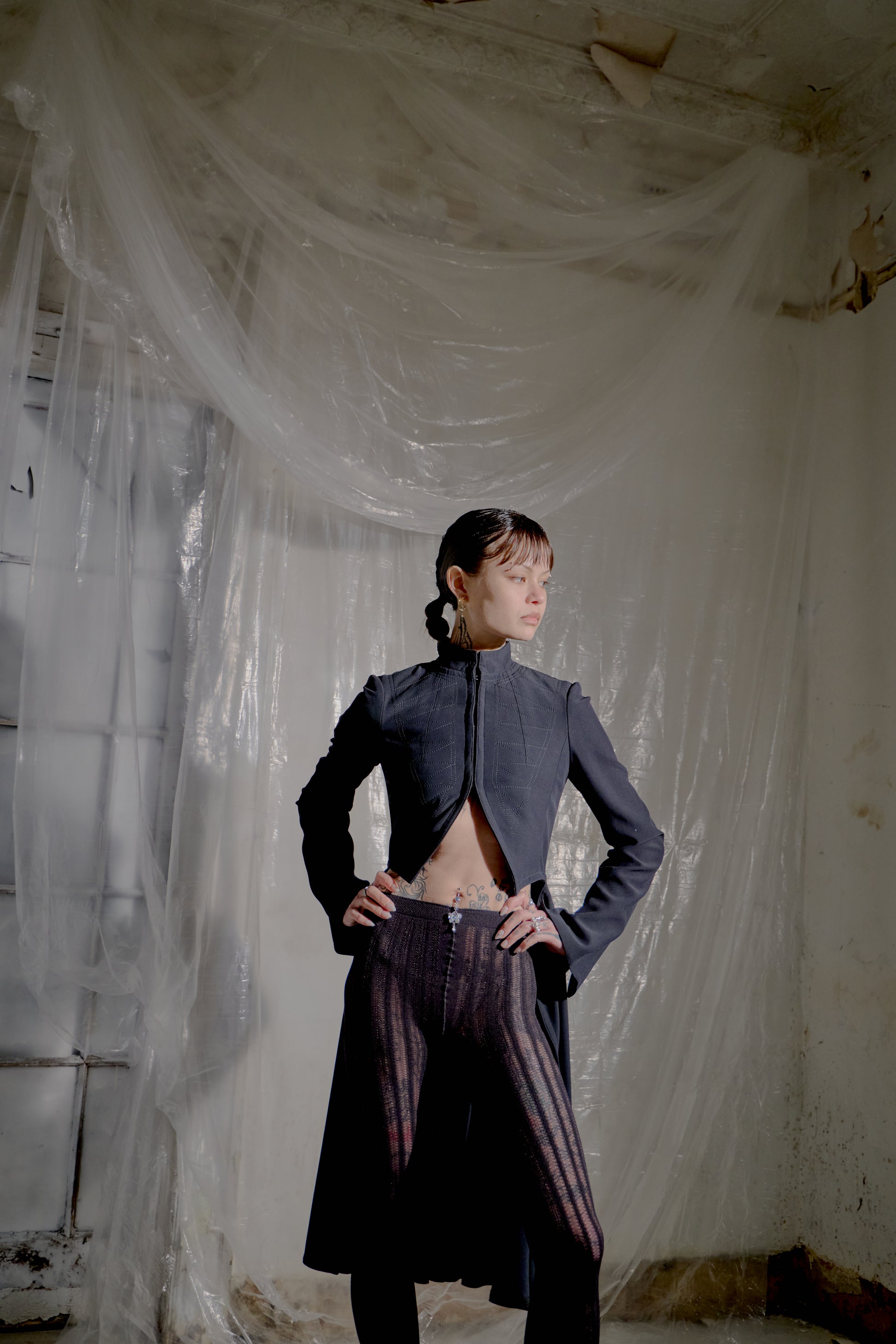 Person wearing a dark Alexander McQueen coat and striped pants standing in a room with plastic sheeting.