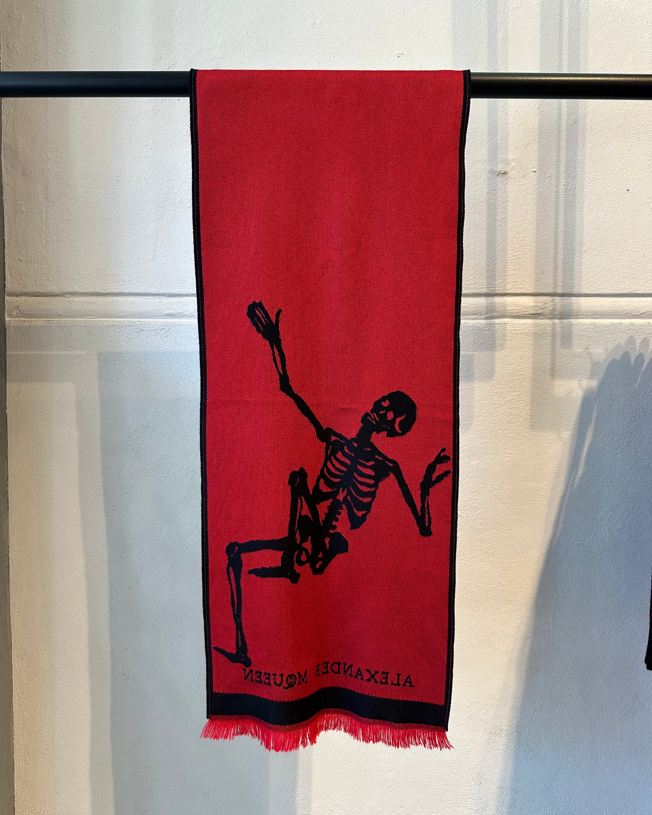 Black Alexander McQueen scarf with red skeleton design hanging on a rack against a light-colored wall.