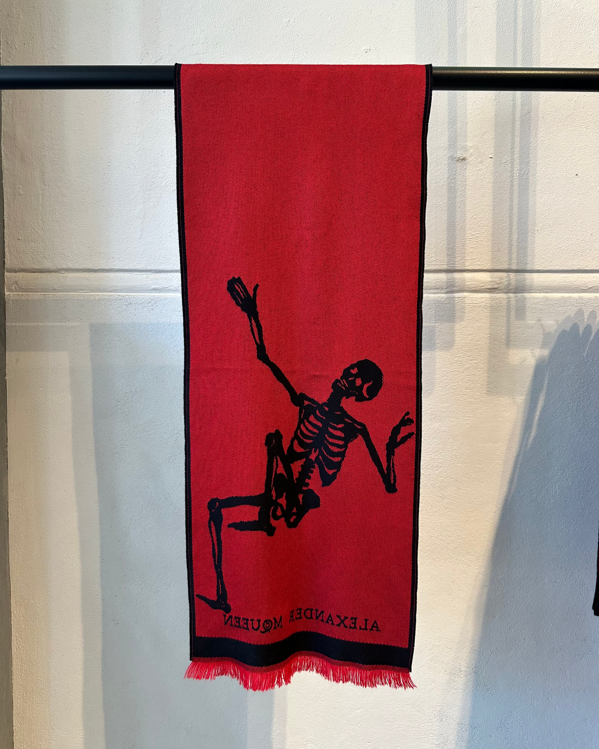 Red Alexander McQueen scarf with black skeleton design hanging on a rack against a white curtain background