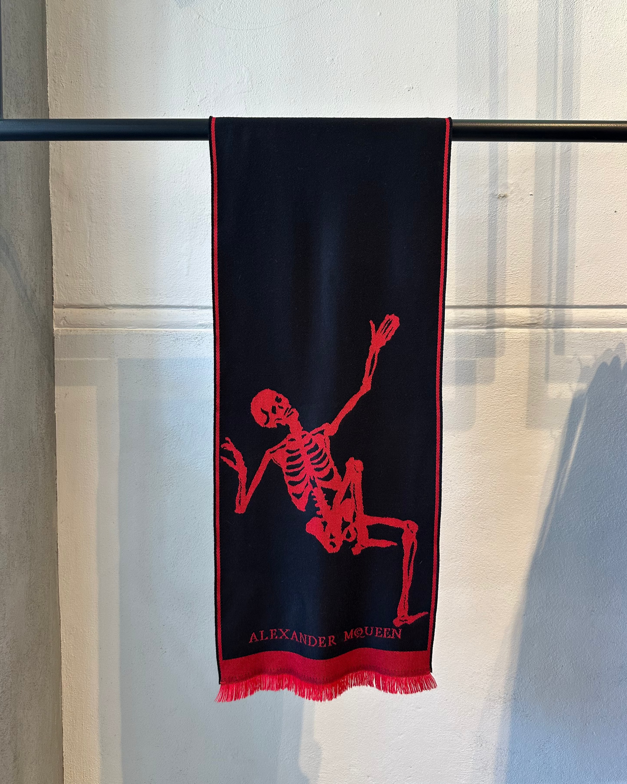 Black Alexander McQueen scarf with red skeleton design hanging on a rack against a light-colored wall.