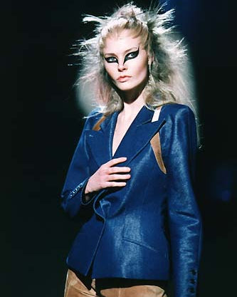 A model on a dimly lit runway wears a blue Alexander McQueen blazer with angular shoulders and brown pants. 