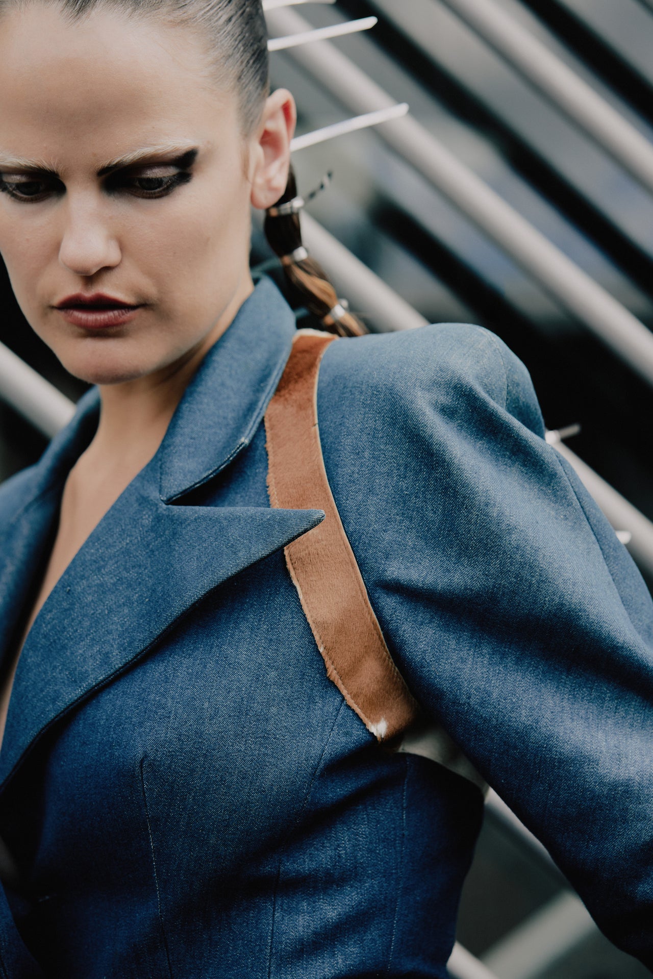 A woman in a blue Alexander McQueen blazer with structured shoulders and a brown strap stands against diagonal metal bars. She has bold eye makeup and an intense expression.