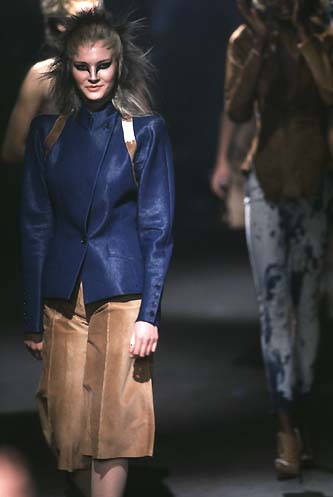 Model on runway in a structured blue Alexander McQueen jacket, tan skirt, and bold eye makeup. Hair is styled with volume, creating an avant-garde look.