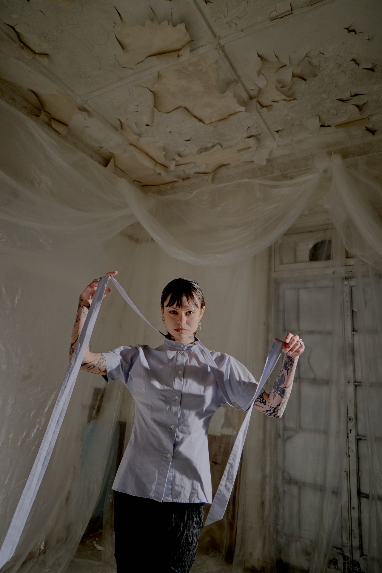 Person holding a ribbon from an Alexander McQueen shirt in an abandoned room with peeling paint.