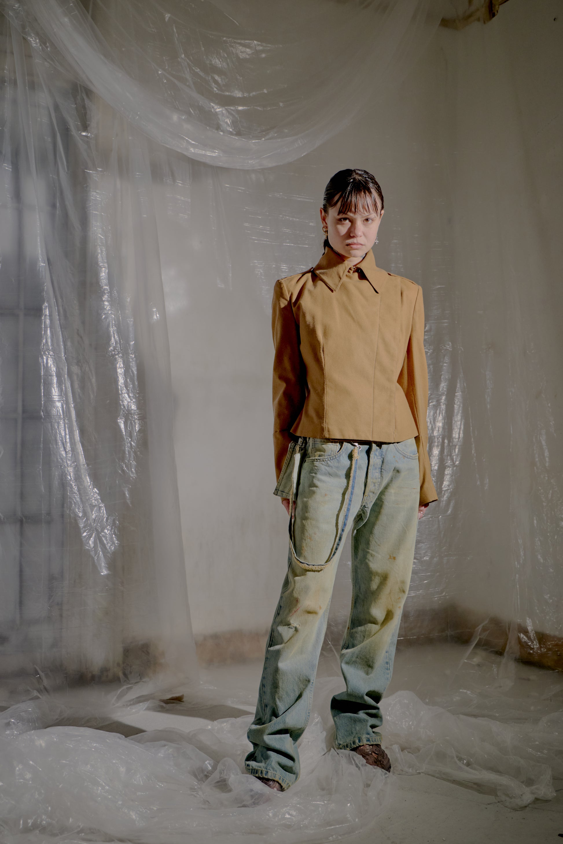 Person wearing a brown Alexander McQueen jacket and denim pants standing in a room with plastic sheeting.