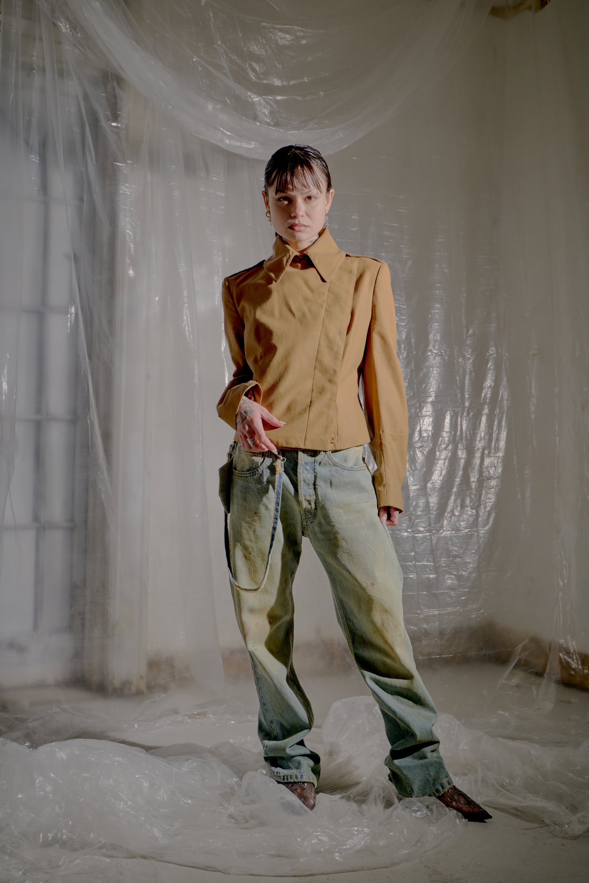 Person wearing a brown Alexander McQueen jacket and denim pants standing in a room with white plastic sheeting.