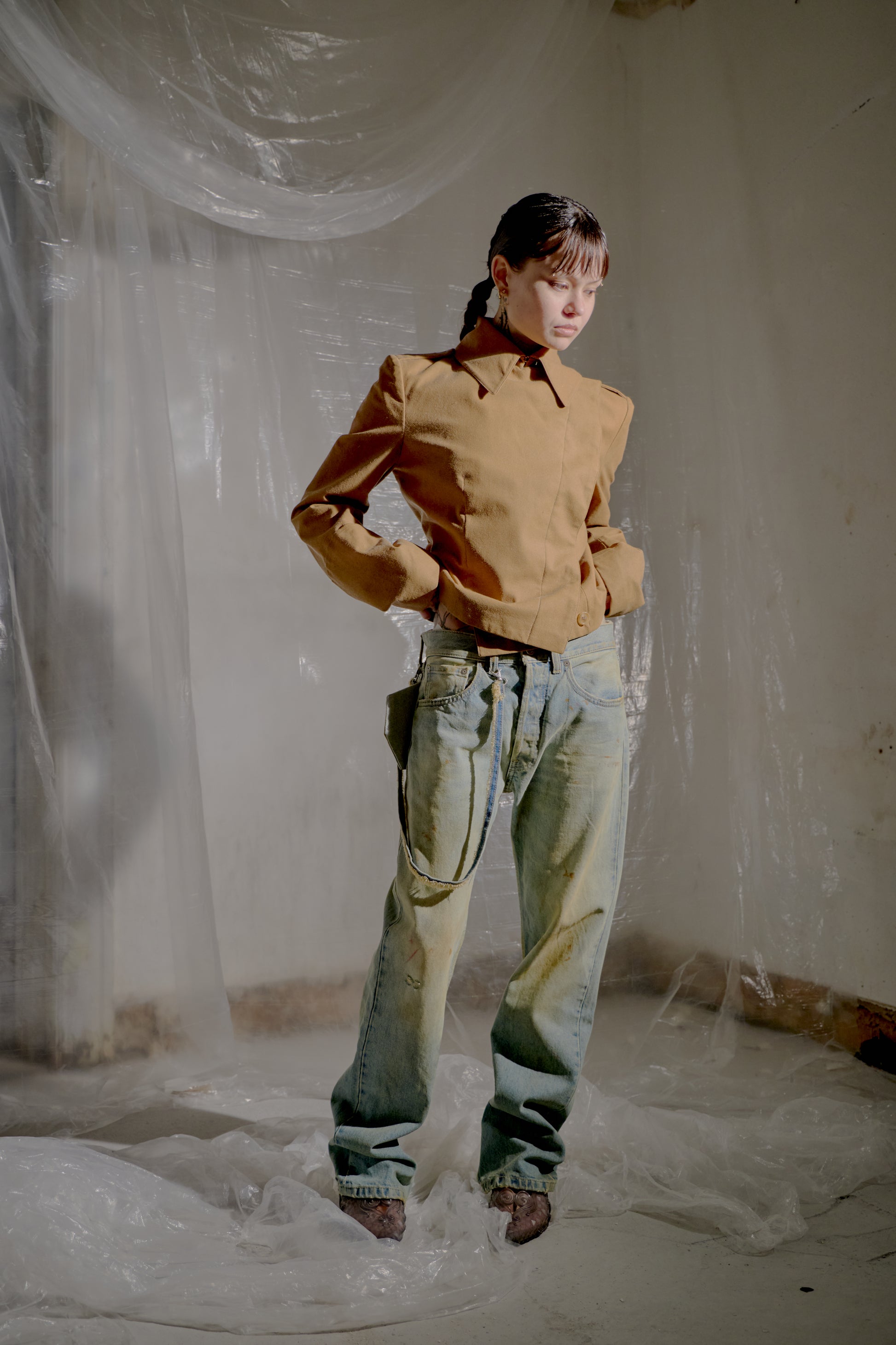 Person wearing a brown Alexander McQueen jacket and denim pants standing in a room with white plastic sheeting.