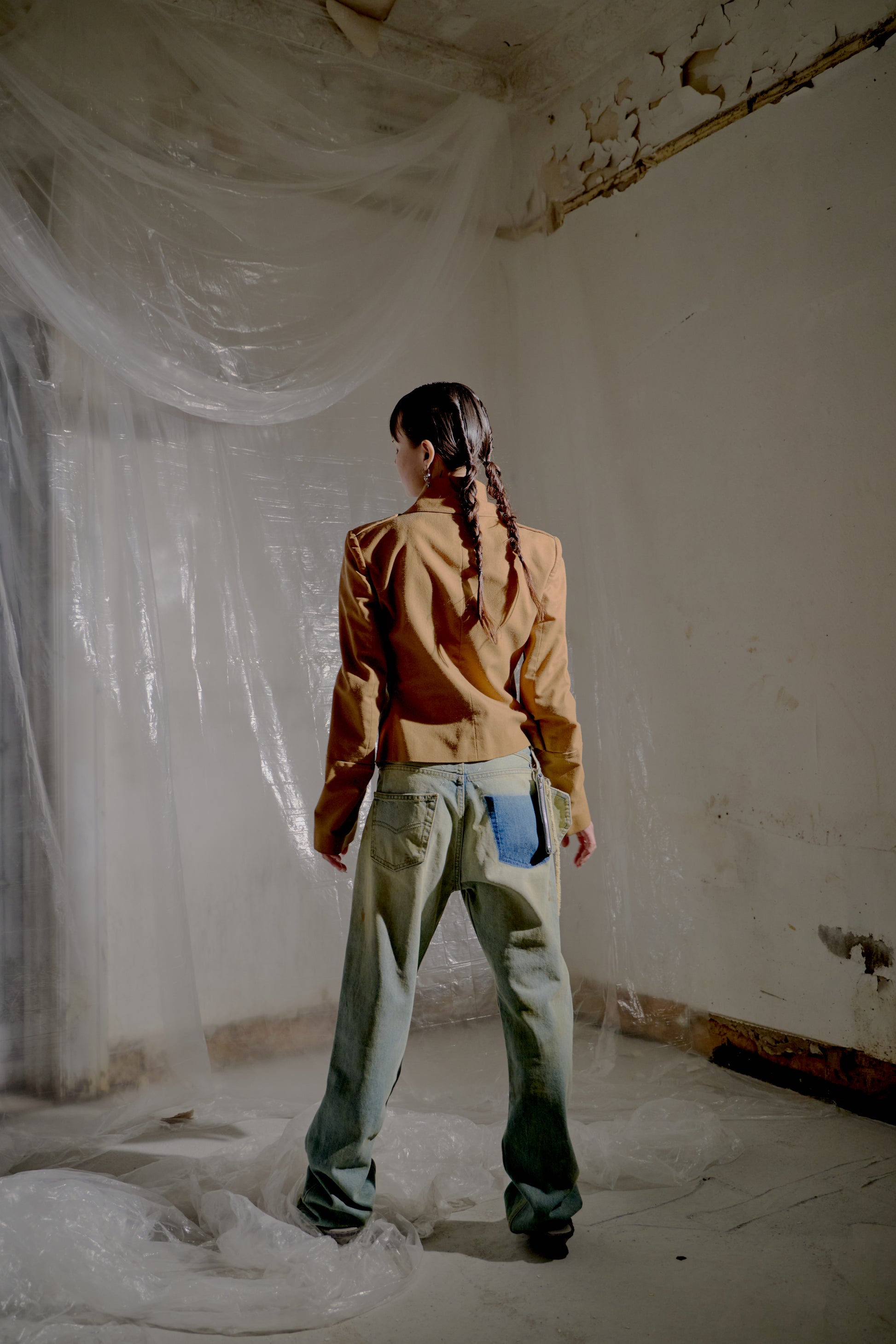 Person with their back to the camera wearing a brown Alexander McQueen jacket and denim pants standing in a room with white plastic sheeting.