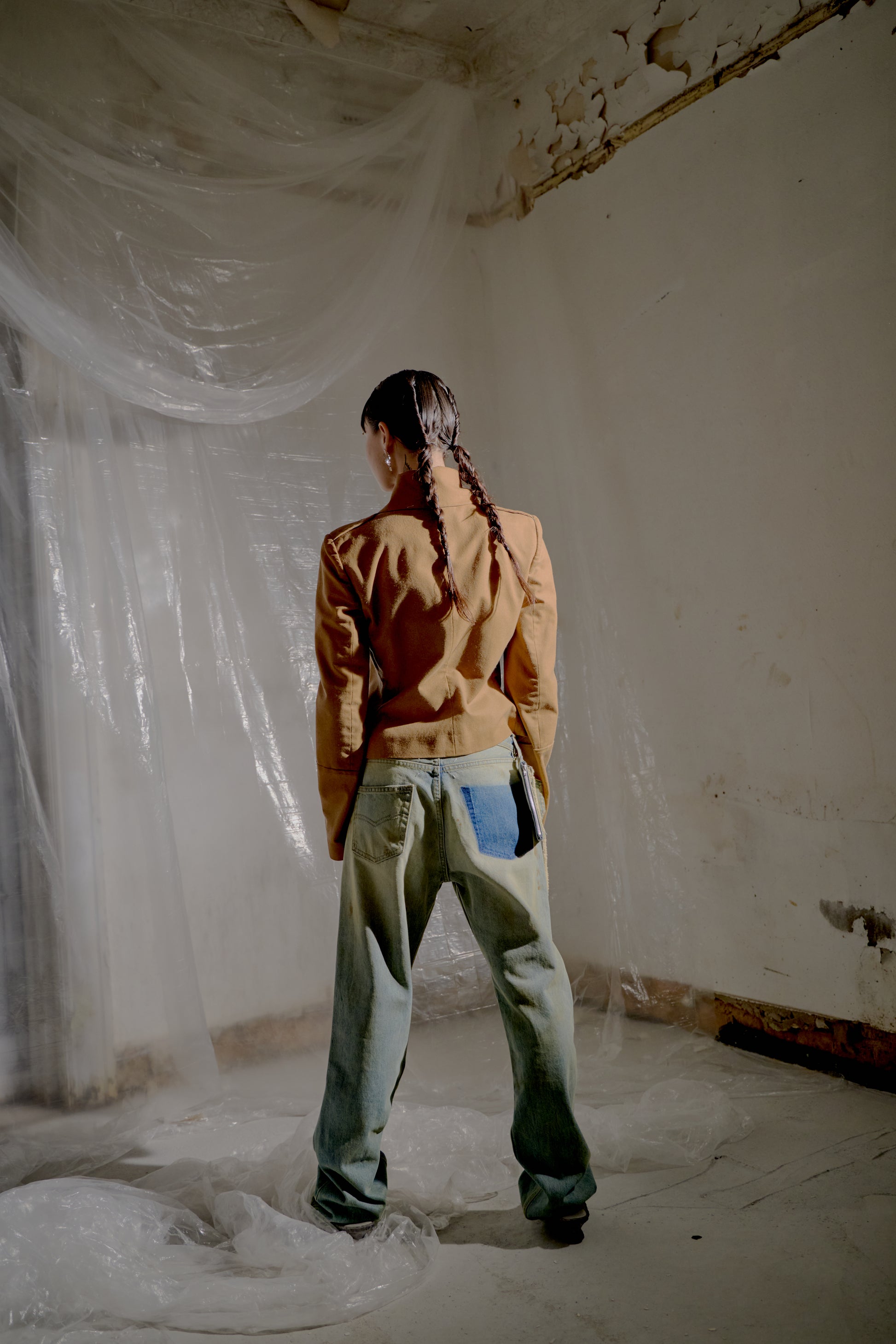 Person standing with their back to the camera wearing a brown Alexander McQueen jacket and denim pants standing in a room with white plastic sheeting.