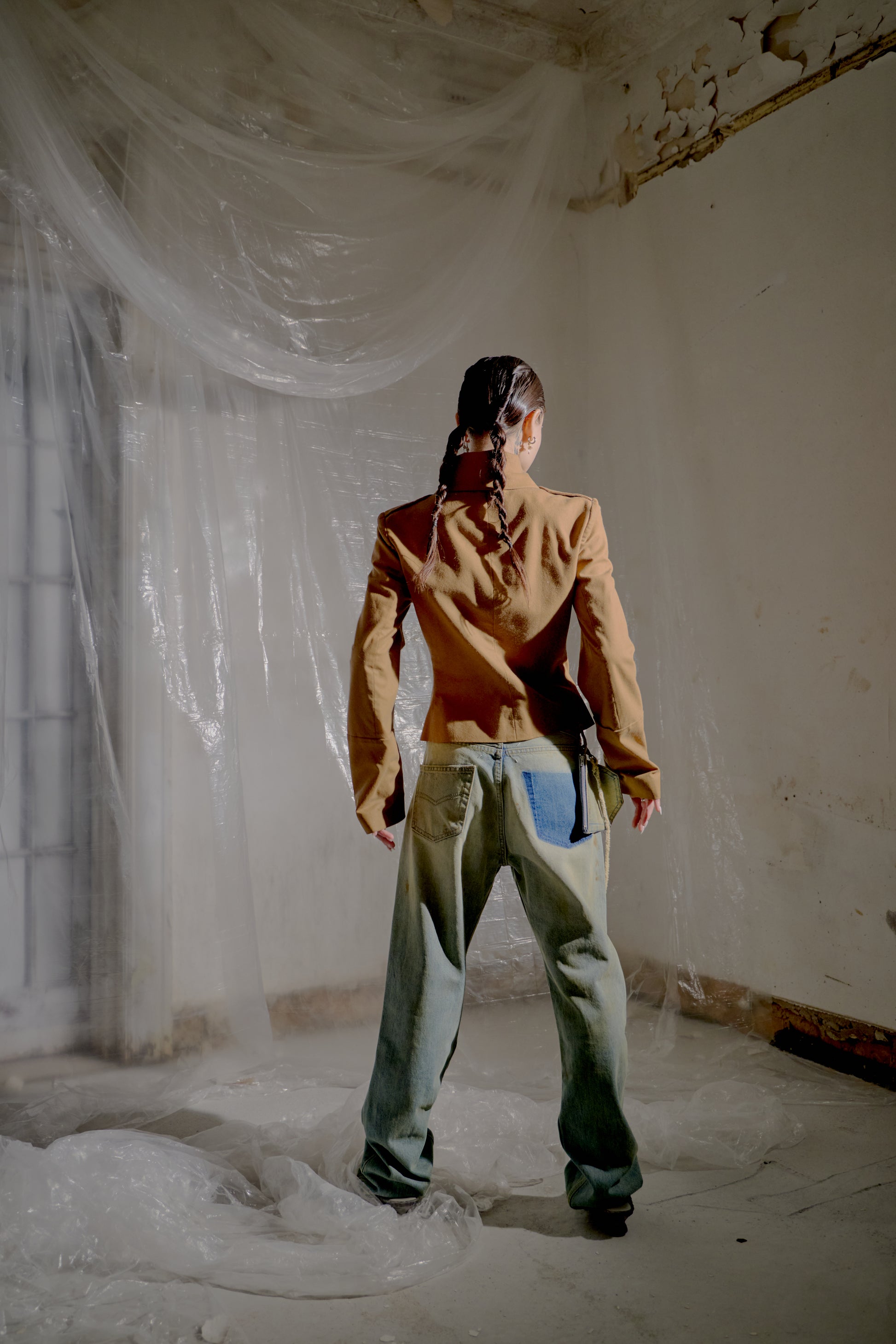 Person wearing a brown Alexander McQueen jacket and denim pants standing in a room with white plastic sheeting.