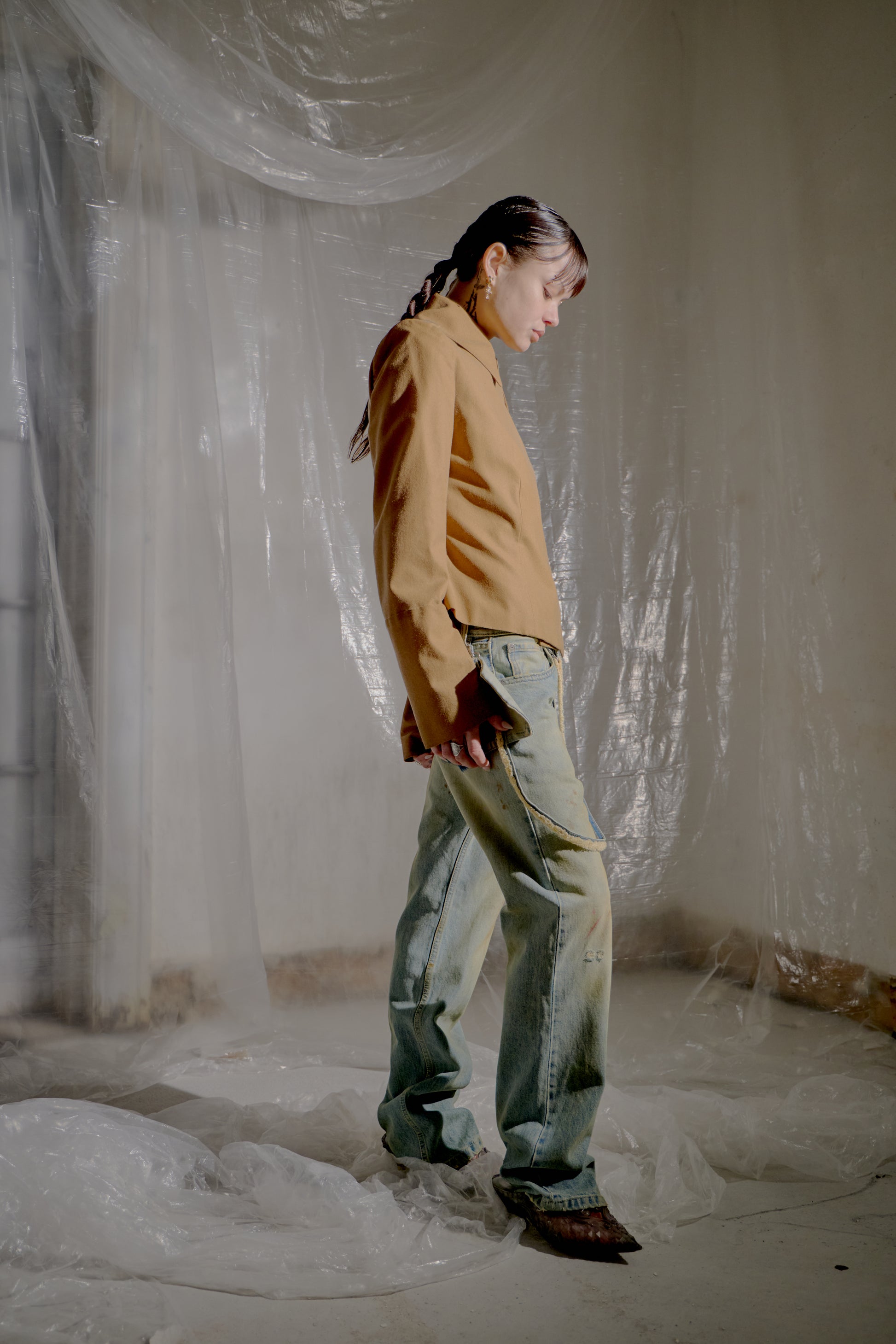 Person wearing a brown Alexander McQueen jacket and denim pants standing in a room with white plastic sheeting.