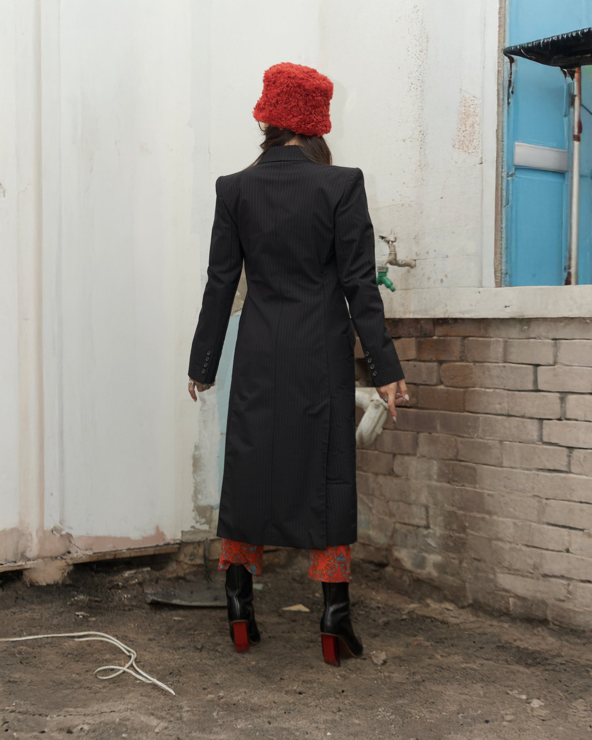 Person wearing a long black Alexander McQueen coat and red hat standing near a wall with a window.