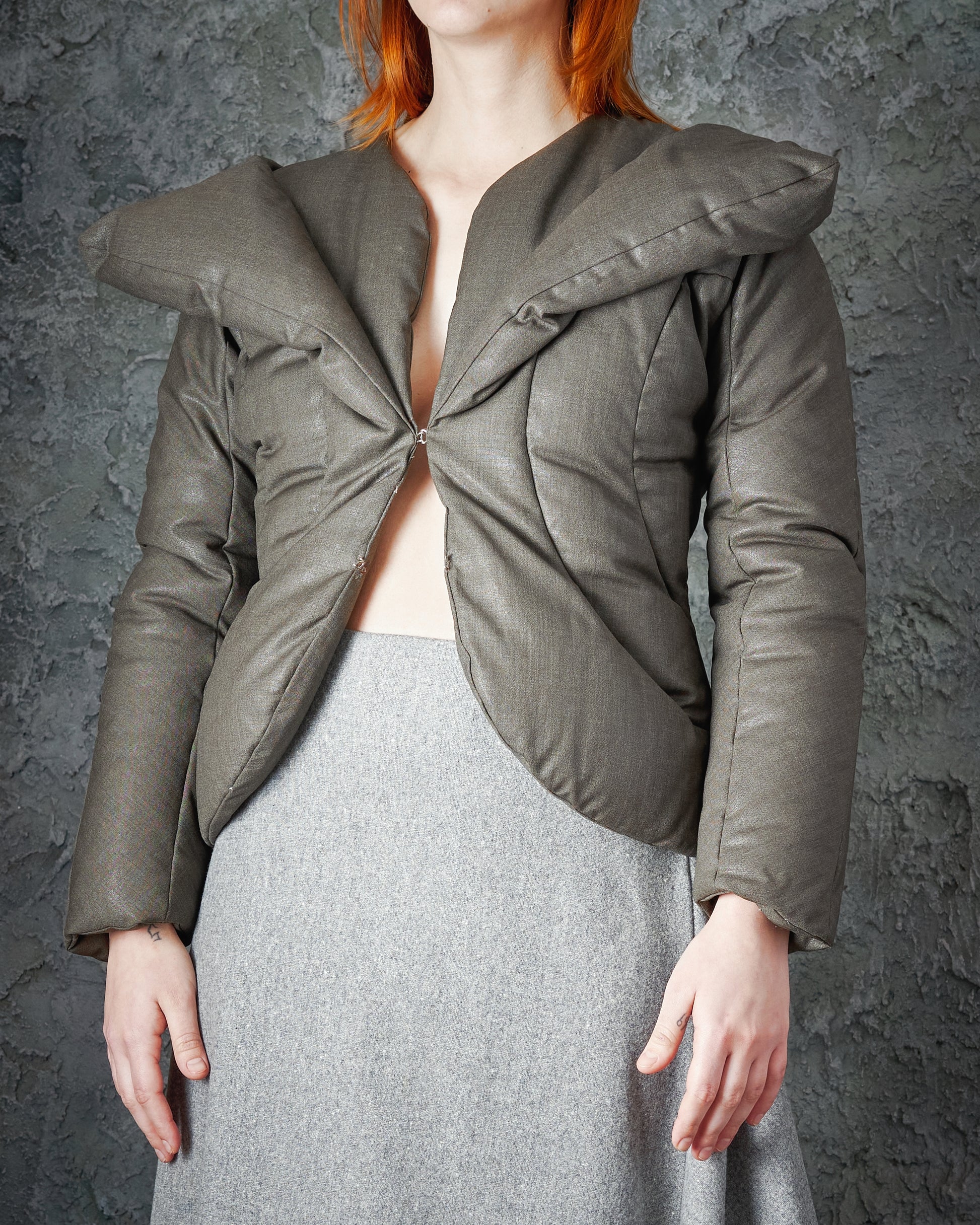 A person wearing a uniquely structured green Alexander McQueen jacket with exaggerated shoulders, paired with a light grey skirt. The background is textured and dark.