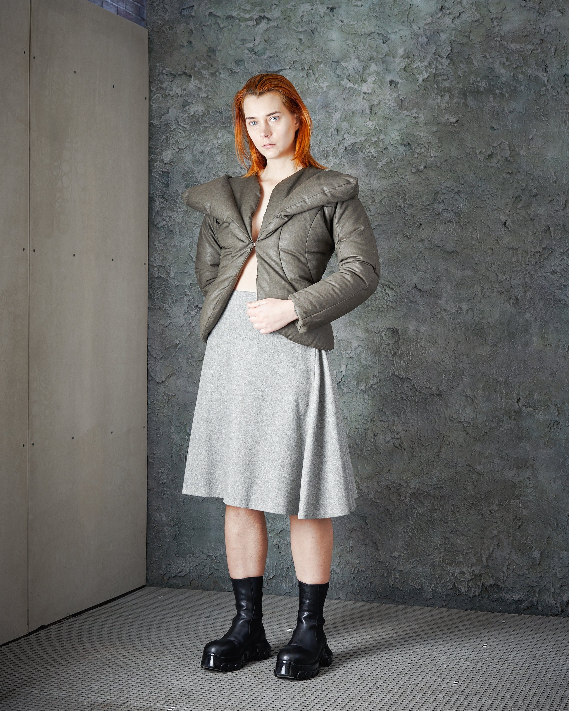 A person with red hair stands in a modern space, wearing an open olive Alexander McQueen jacket, grey skirt, and black boots. The atmosphere is edgy and stylish.