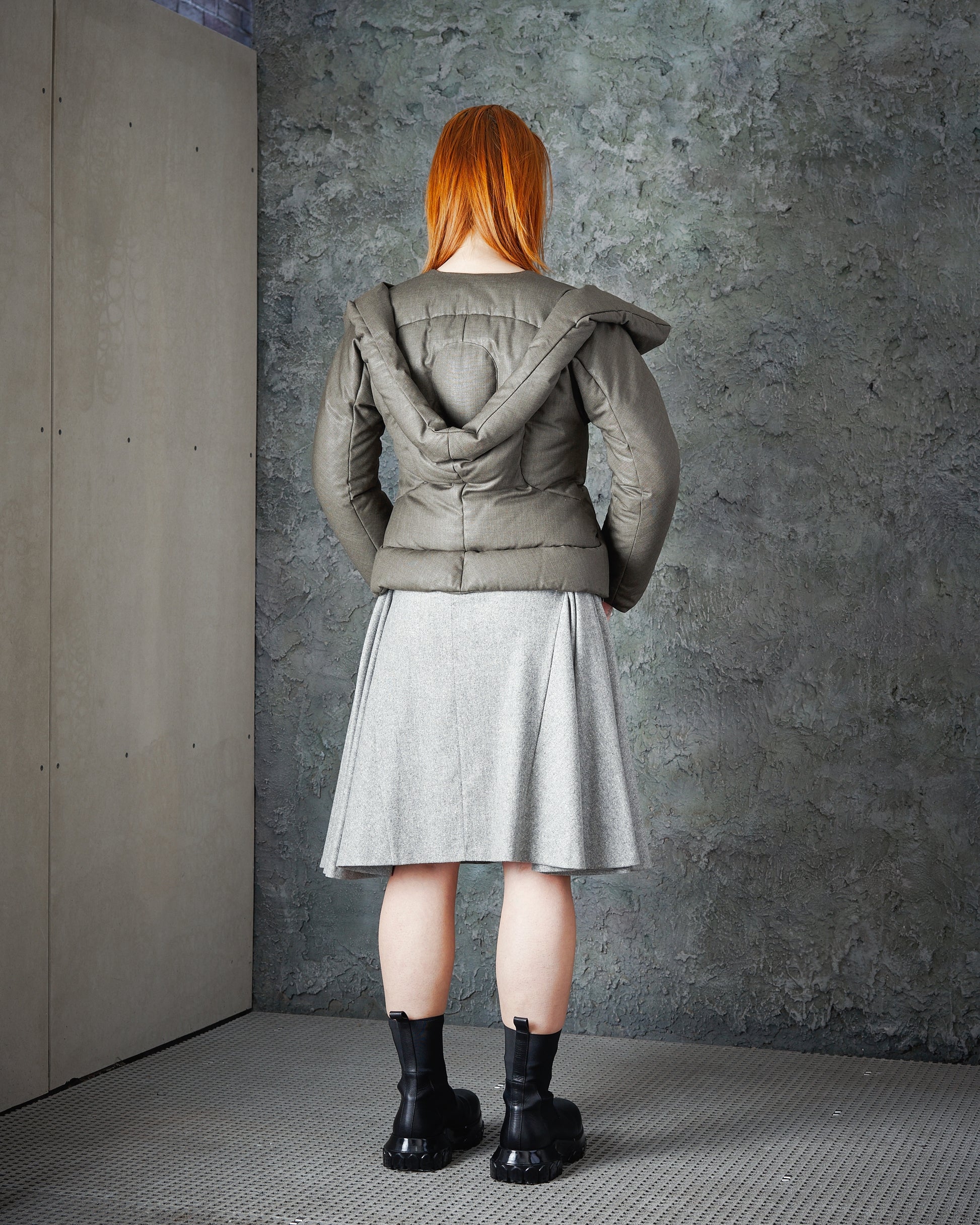 A person with long red hair stands facing a textured wall, wearing a green Alexander McQueen puffer jacket, light grey skirt, and black boots, conveying a neutral tone.