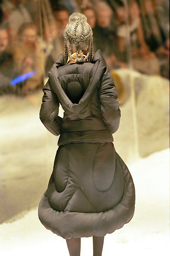 A model with braided hair walks on a fashion runway wearing a unique green, quilted Alexander McQueen coat with a hood. The audience is blurred in the background.