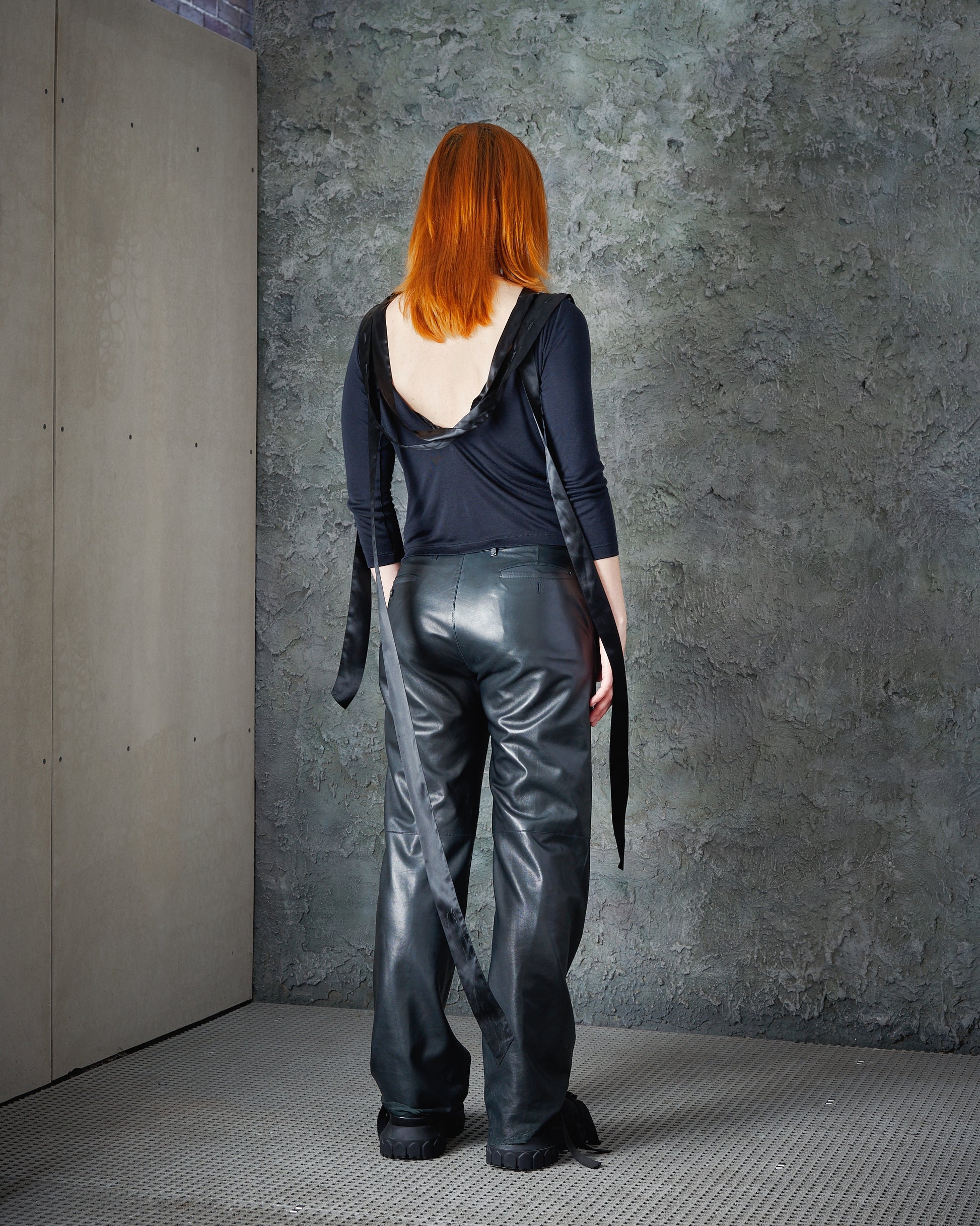 A person with red hair is seen from behind, wearing a dark, backless Alexander McQueen top and loose leather pants.