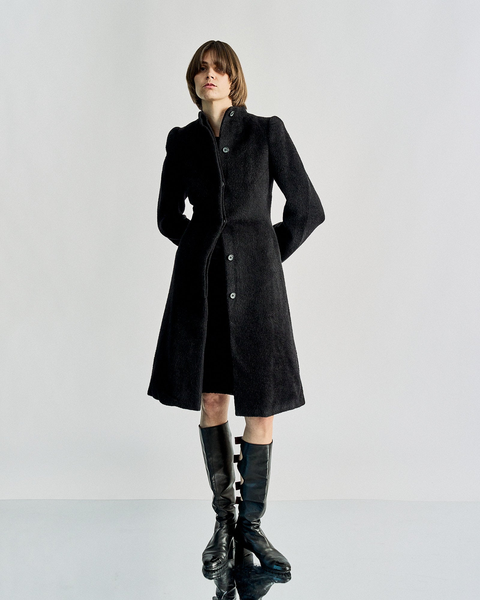 A person is wearing a buttoned, black textured Alexander McQueen coat and knee-high glossy black boots, standing against a plain light grey background, exuding a sophisticated and confident mood.