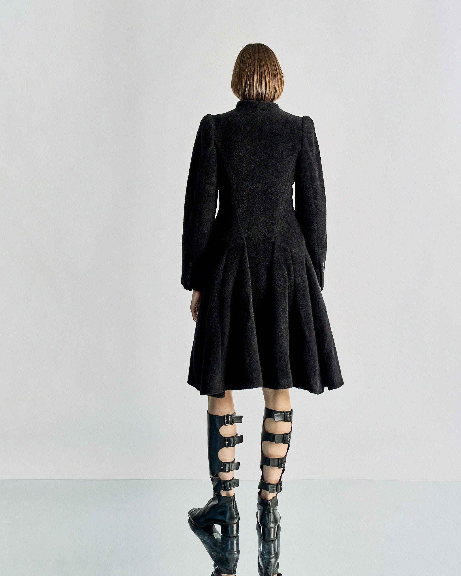 A person with a bob haircut is wearing a long, black Alexander McQueen coat and knee-high, strappy leather boots. The minimalist background enhances the edgy vibe.