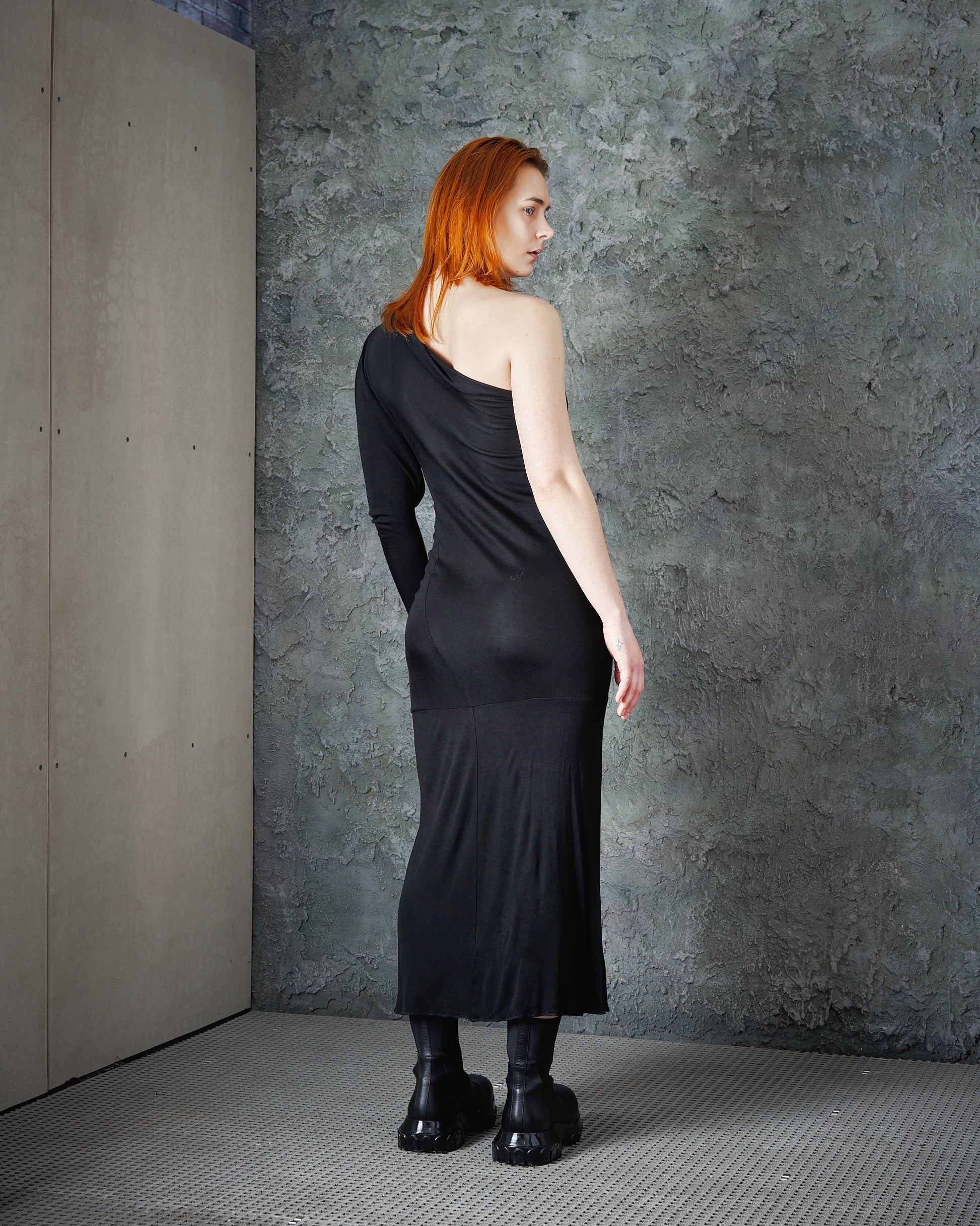 A woman with red hair in a black, one-shoulder Alexander McQueen dress stands against a textured gray wall. She wears black boots