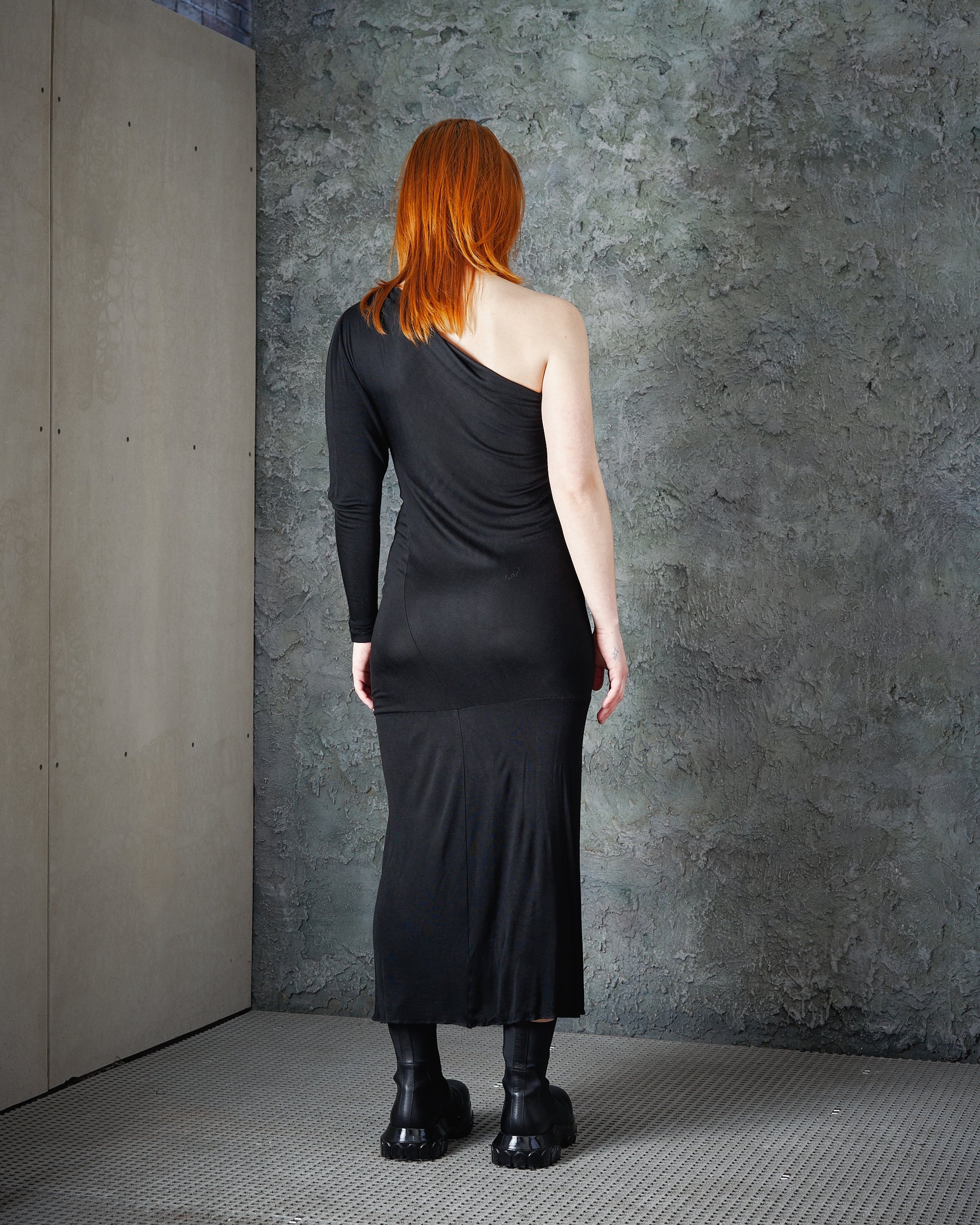 A person with long red hair faces a textured grey wall, wearing a sleek black Alexander McQueen one-sleeve dress and chunky black boots. 