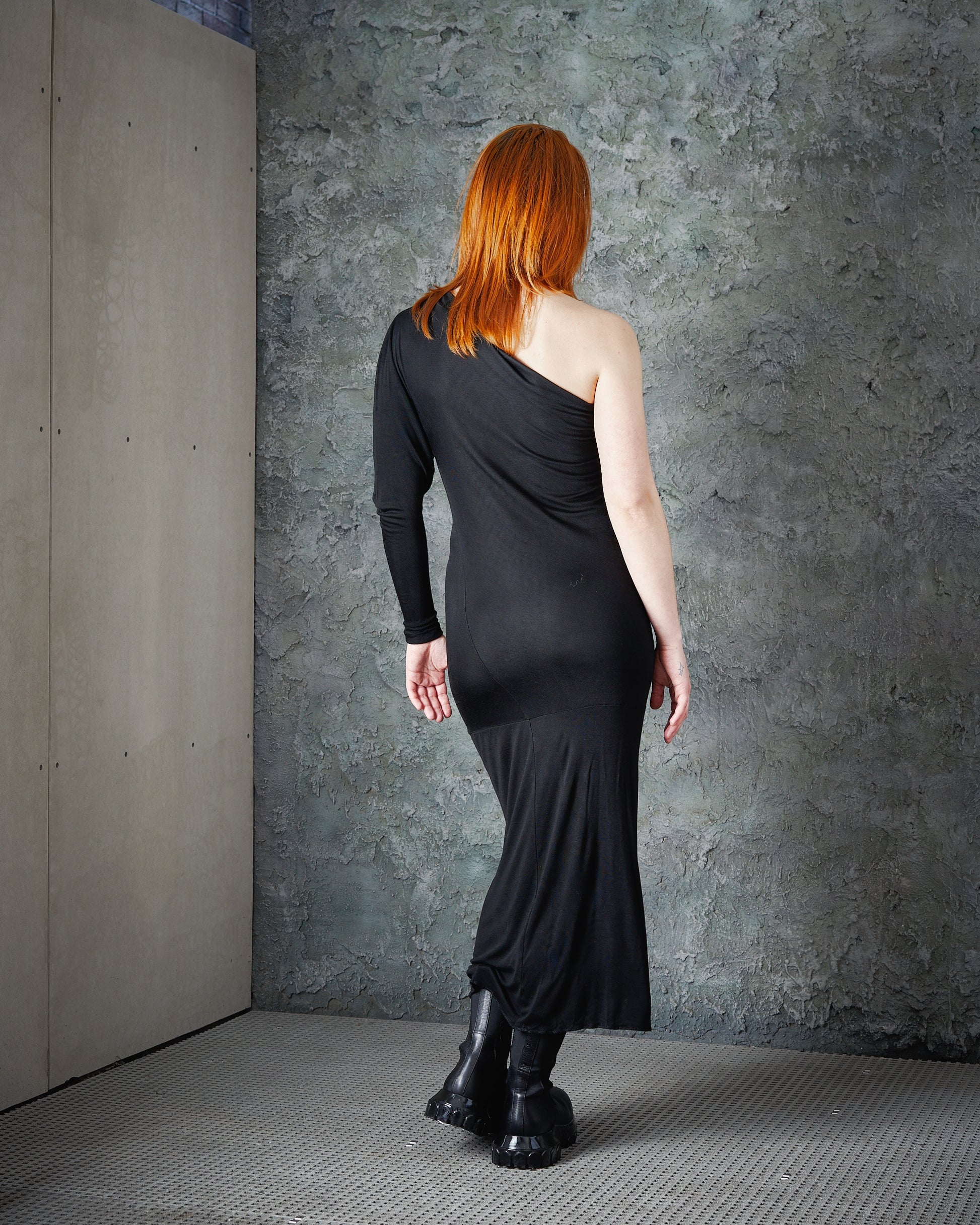 A person with long red hair faces a textured grey wall, wearing a sleek black Alexander McQueen one-sleeve dress and chunky black boots. The tone is bold and edgy.