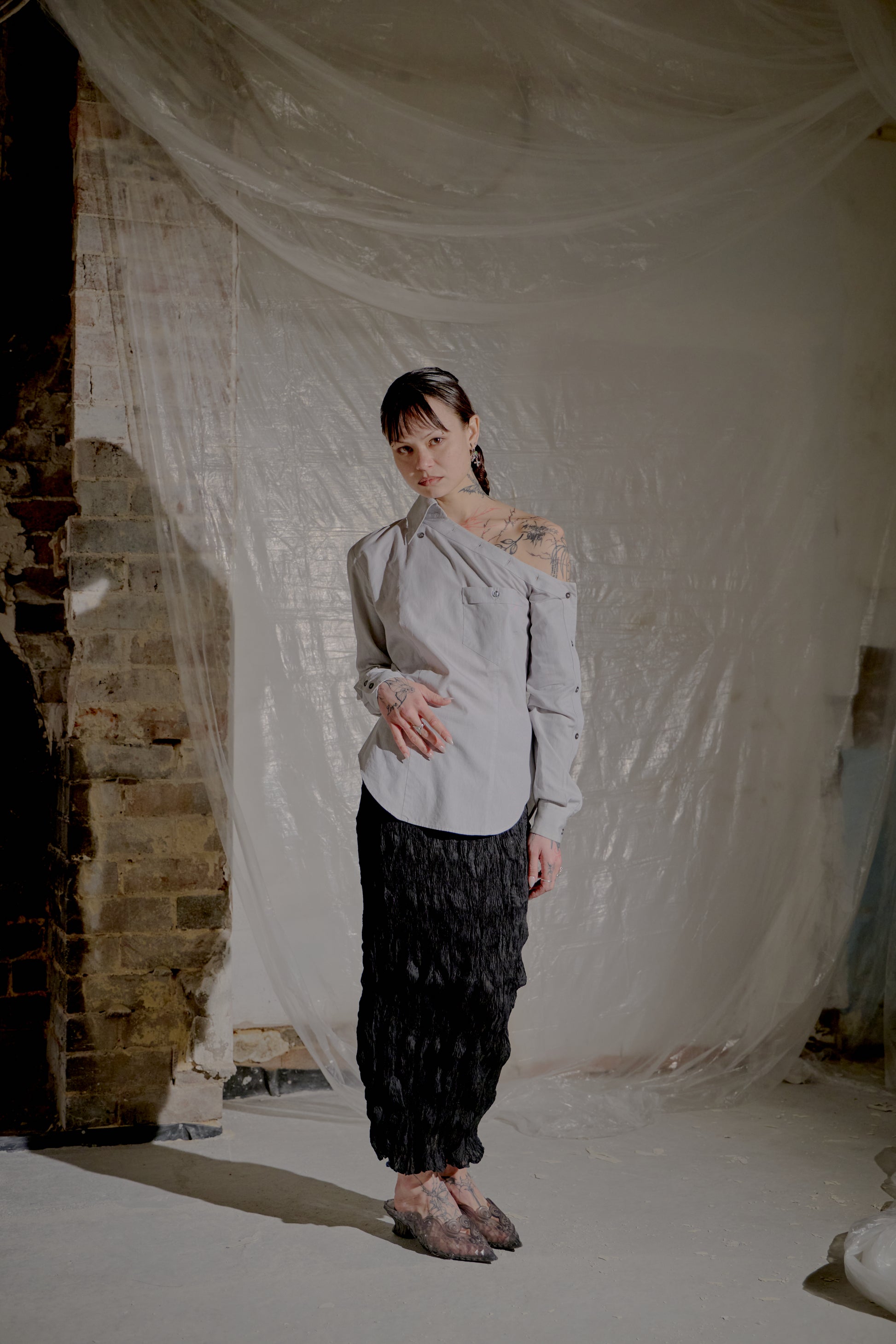 Person wearing a light-coloured Alexander McQueen blouse and dark pants standing in a room with white fabric and brick wall.