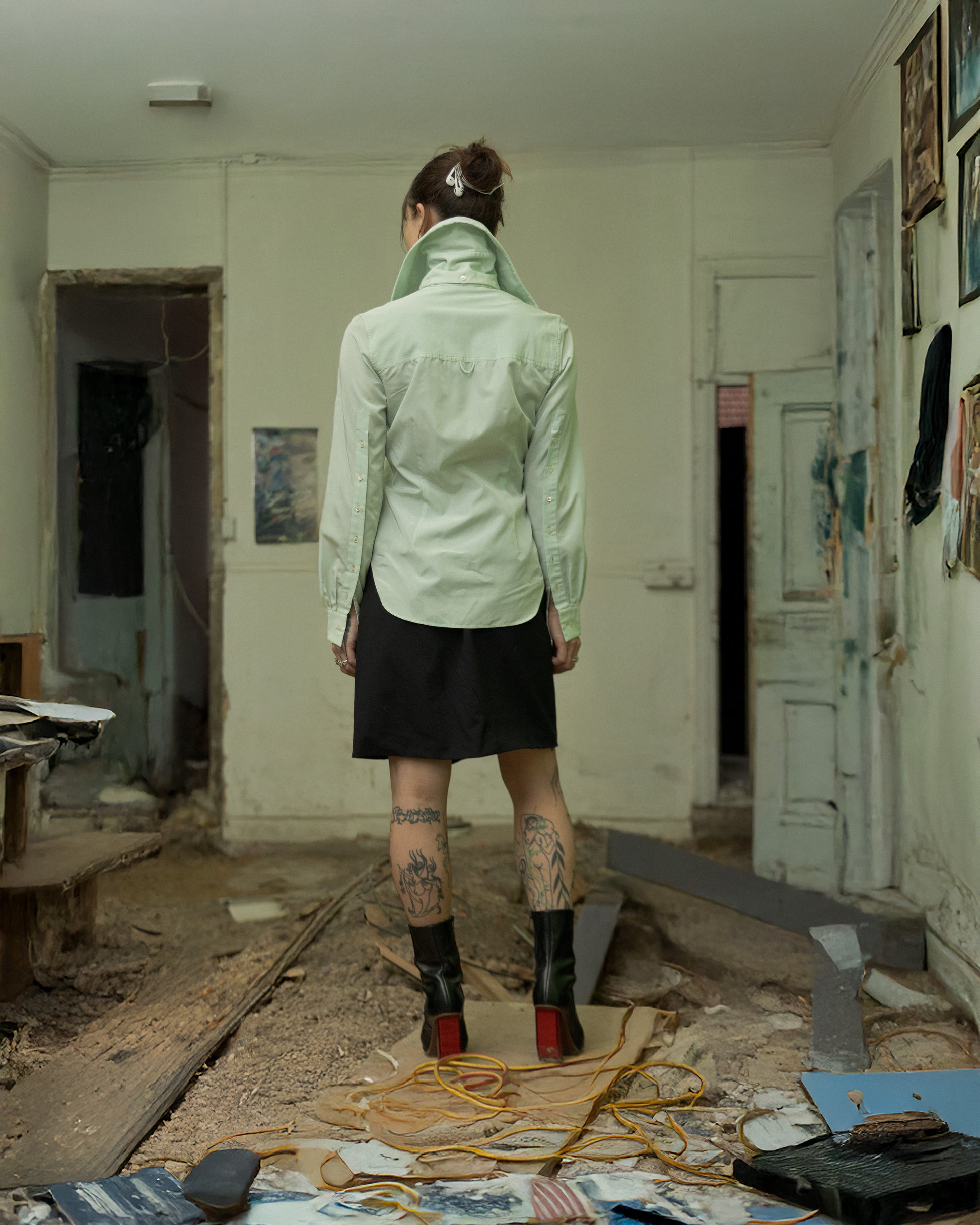 Person wearing a light green Alexander McQueen shirt with a high collar in an abandoned room with debris and artworks.