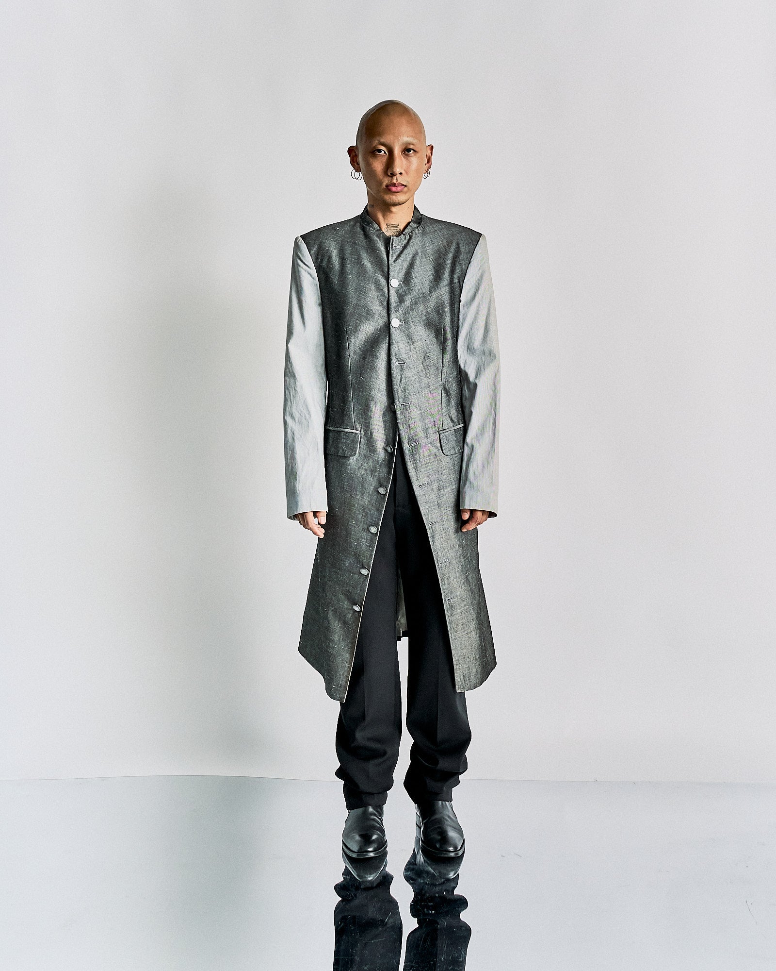 Bald person wearing a metallic grey long Alexander McQueen coat with light grey sleeves, black pants, and boots, standing against a minimalist white background.
