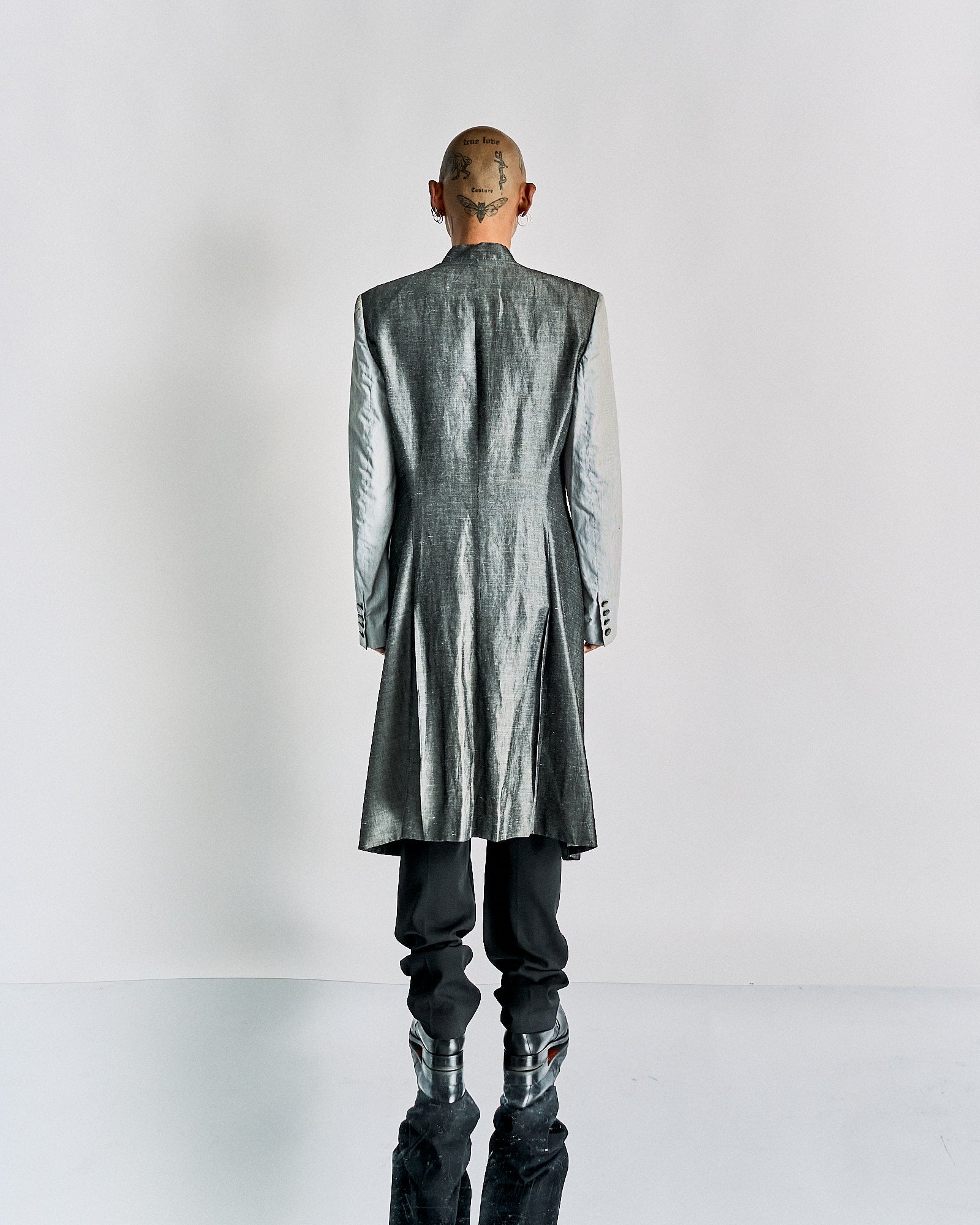A person in a silver long Alexander McQueen coat and black pants stands with their back to the viewer. Their shaved head features tattoos, and they wear metallic boots.