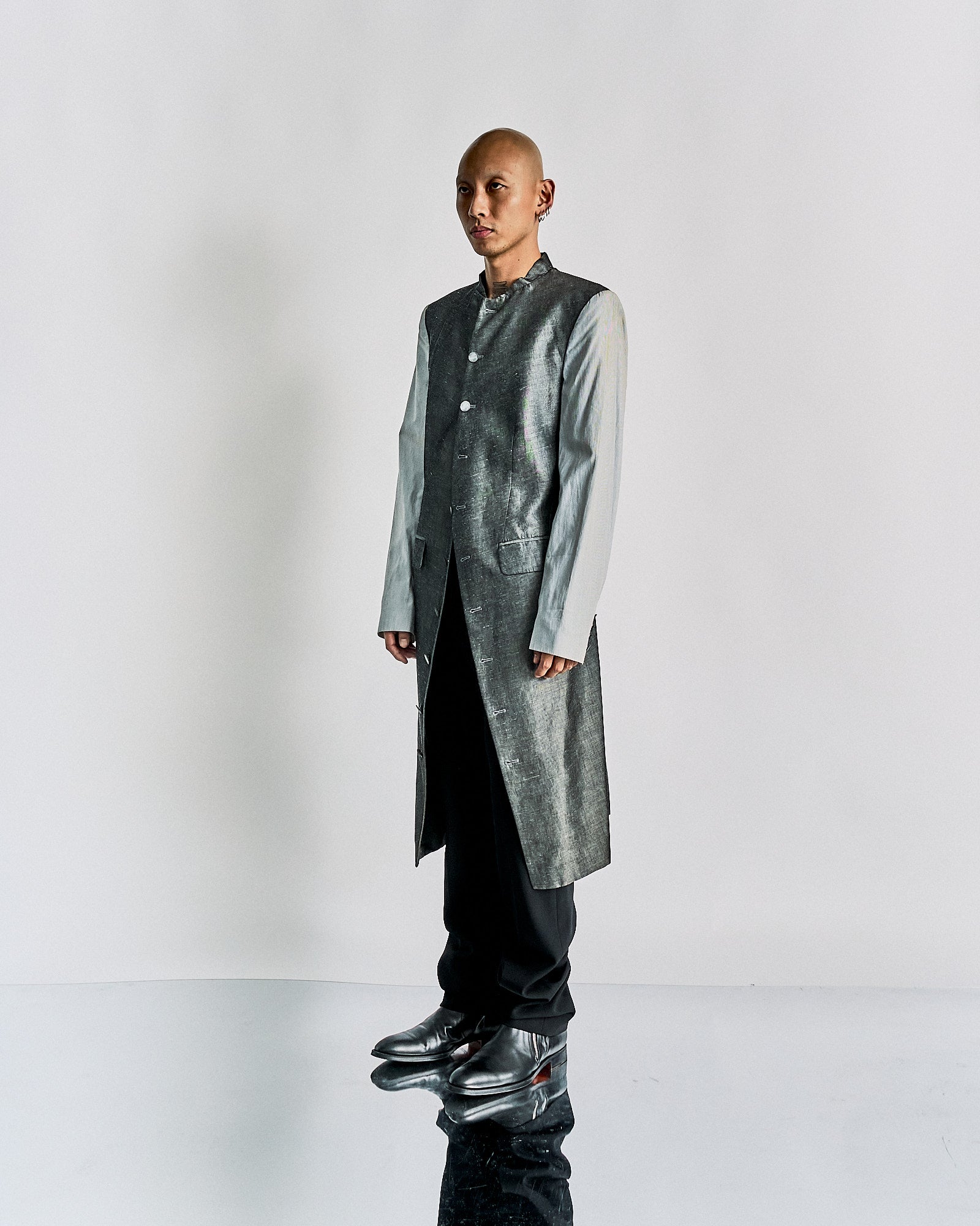 A person stands in a modern long Alexander McQueen coat, blending silver and light gray tones, with black pants and shiny shoes. The setting is minimalist and sleek.