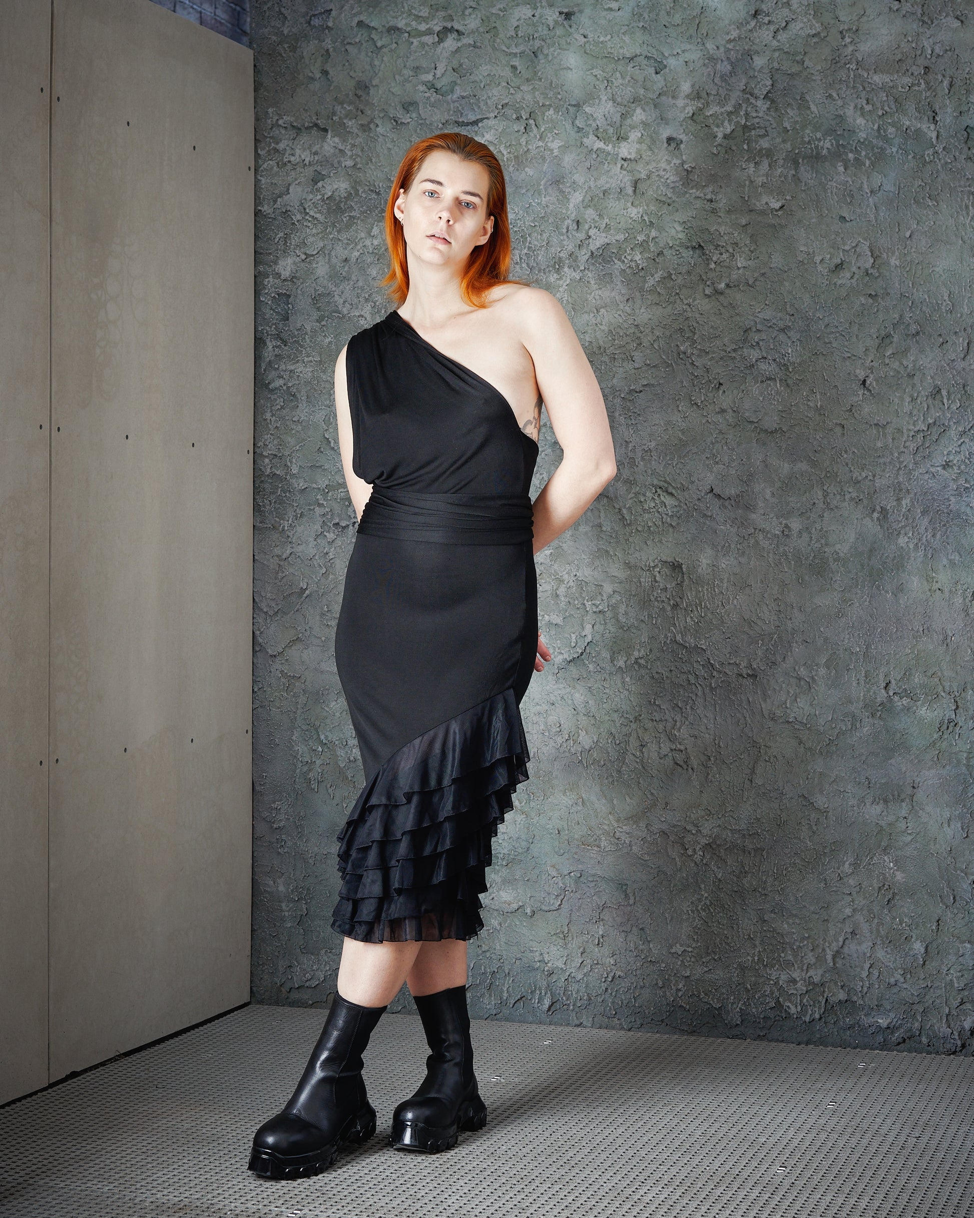 A person with long red hair is wearing a stylish black Alexander McQueen asymmetrical dress and heavy black boots, against a textured grey wall.