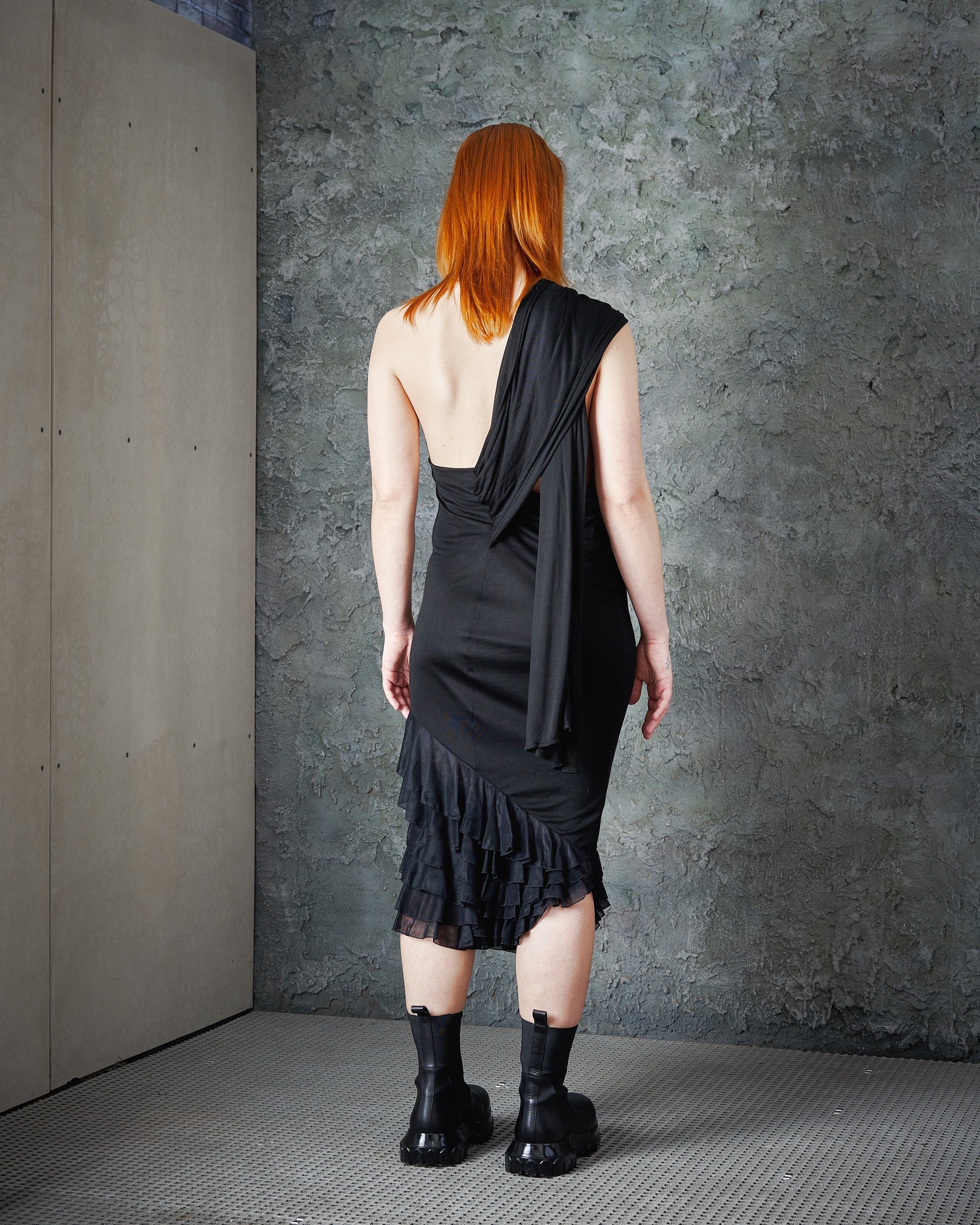 A person with long red hair is seen from behind, wearing a stylish black Alexander McQueen asymmetrical dress and heavy black boots, against a textured grey wall.