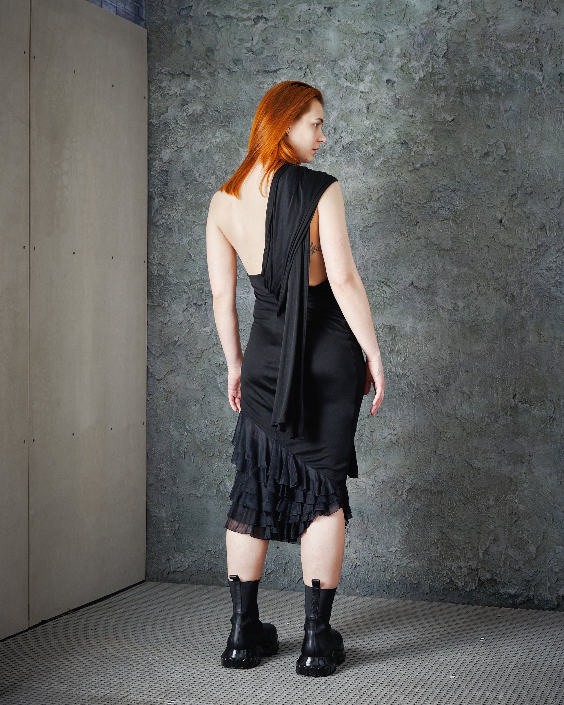 A person with long red hair is seen from behind, wearing a stylish black Alexander McQueen asymmetrical dress and heavy black boots, against a textured grey wall.