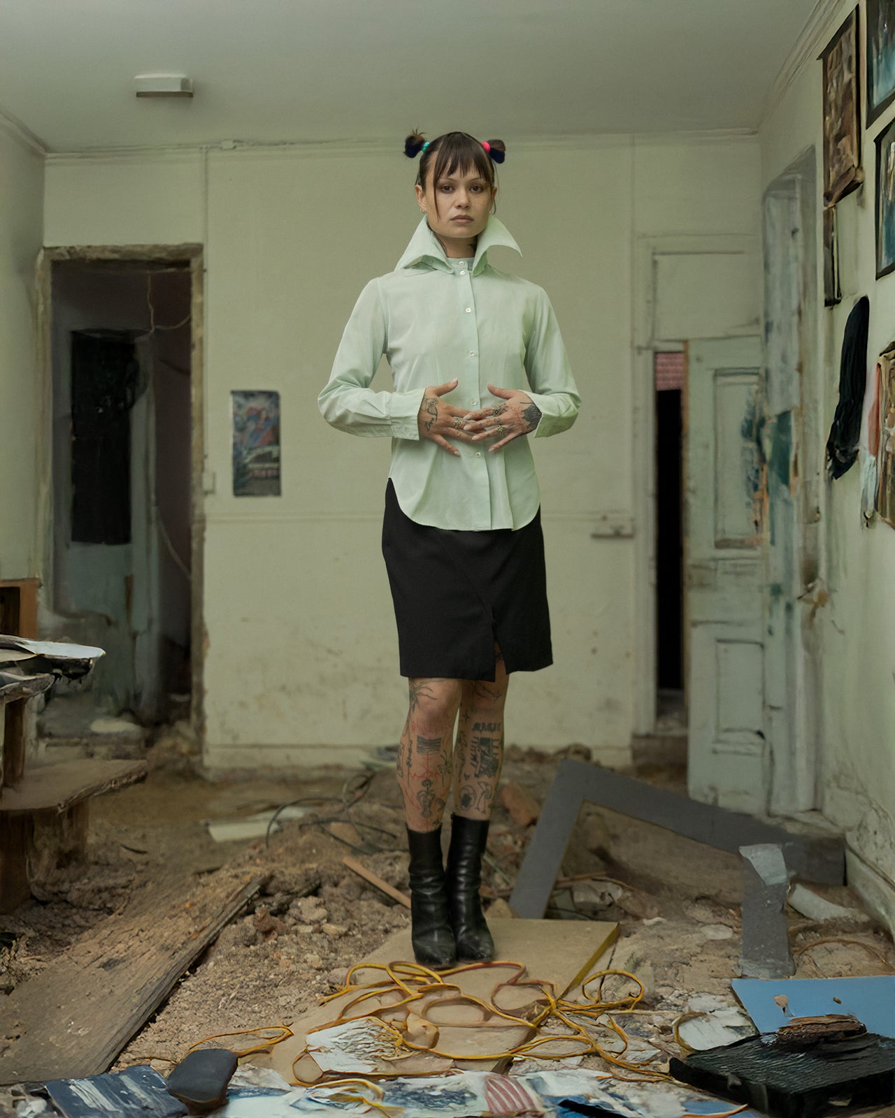 A person stands in a dilapidated room wearing a green Alexander McQueen high collar two-way shirt with a black skirt and black shoes.