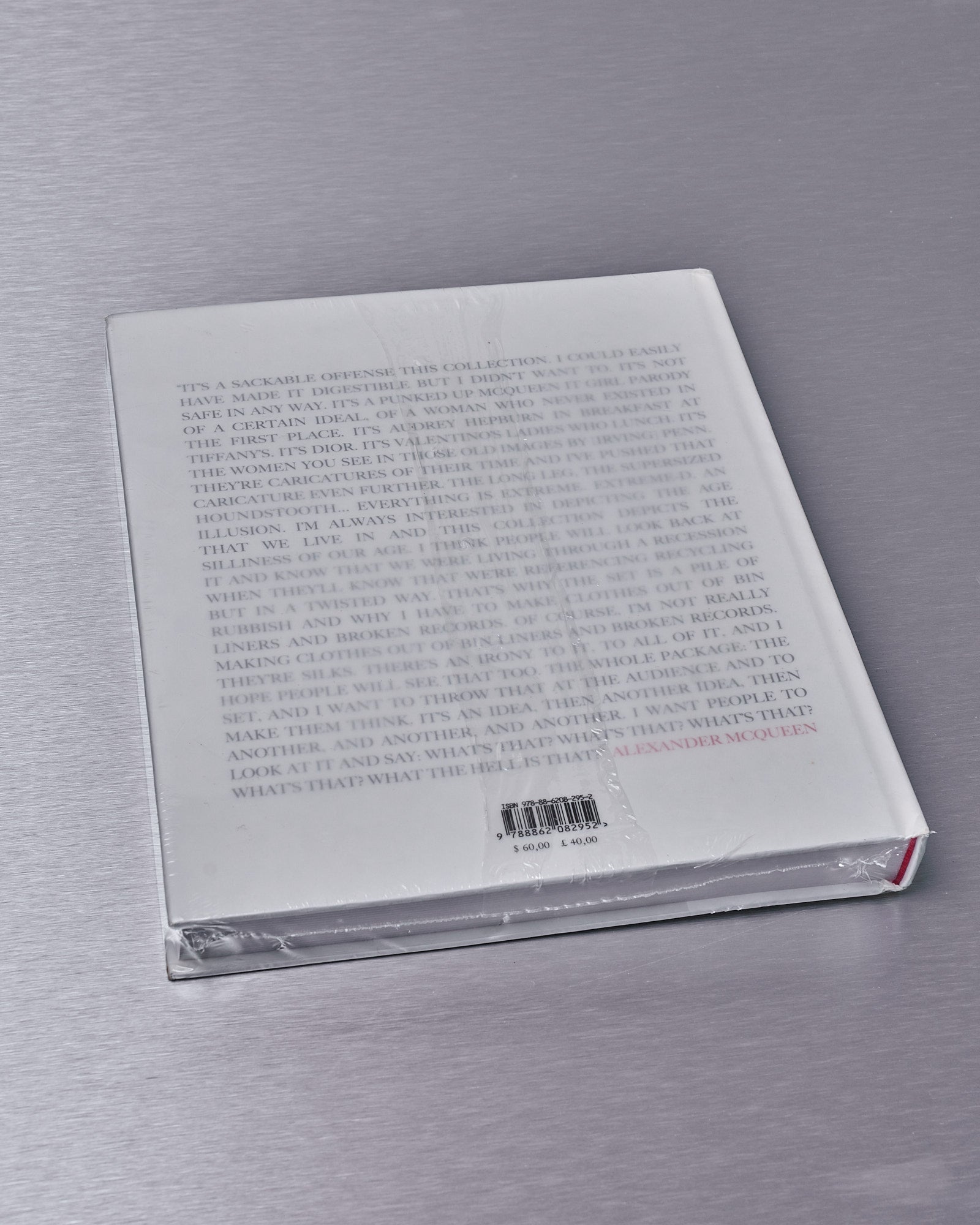 Back cover of an Alexander McQueen book on a metallic surface, wrapped in plastic. It features an extensive text block with black and red text above a barcode.