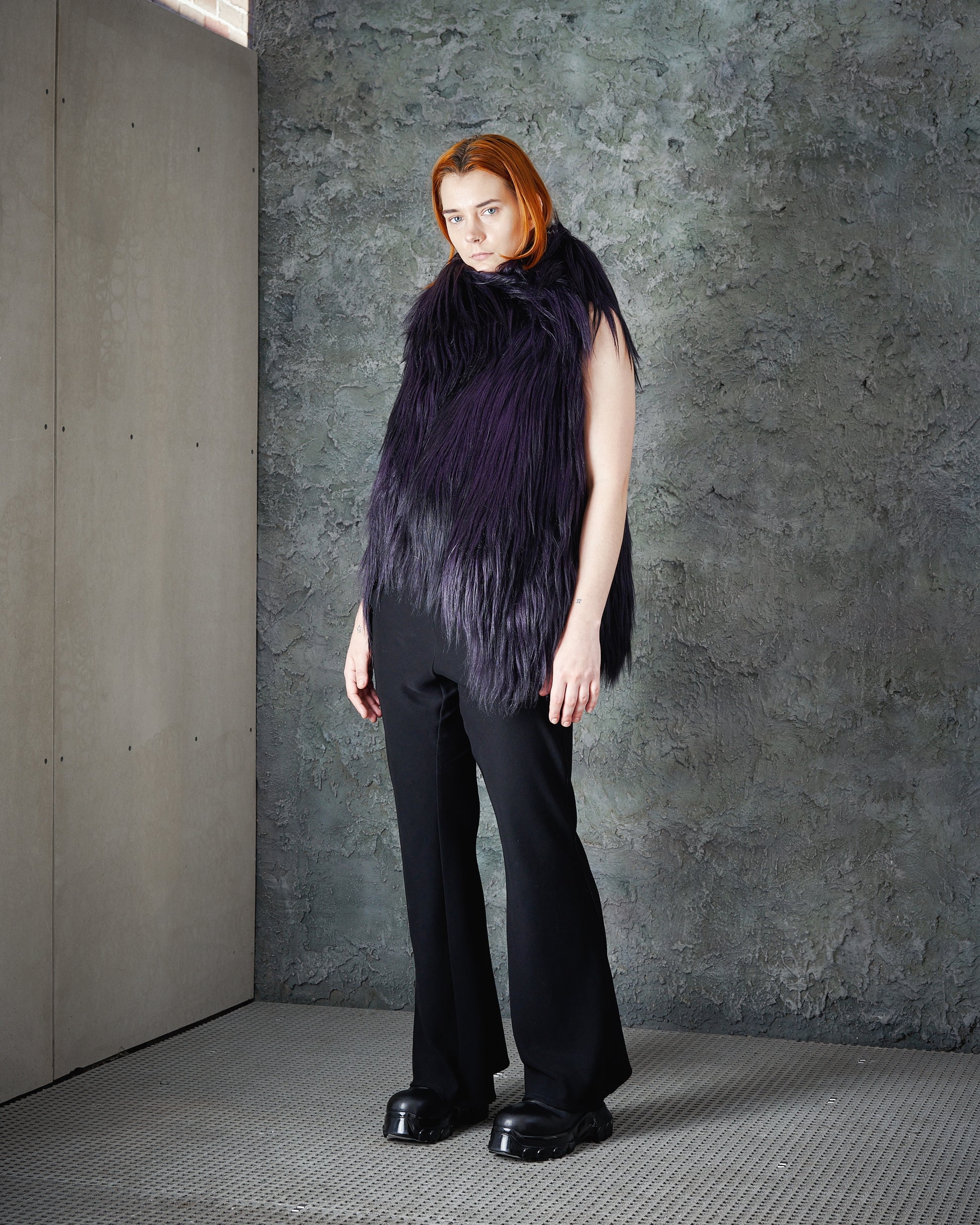 A person stands against a textured gray wall, wearing a purple faux-fur Ann Demeulemeester vest, black pants, and chunky black shoes.