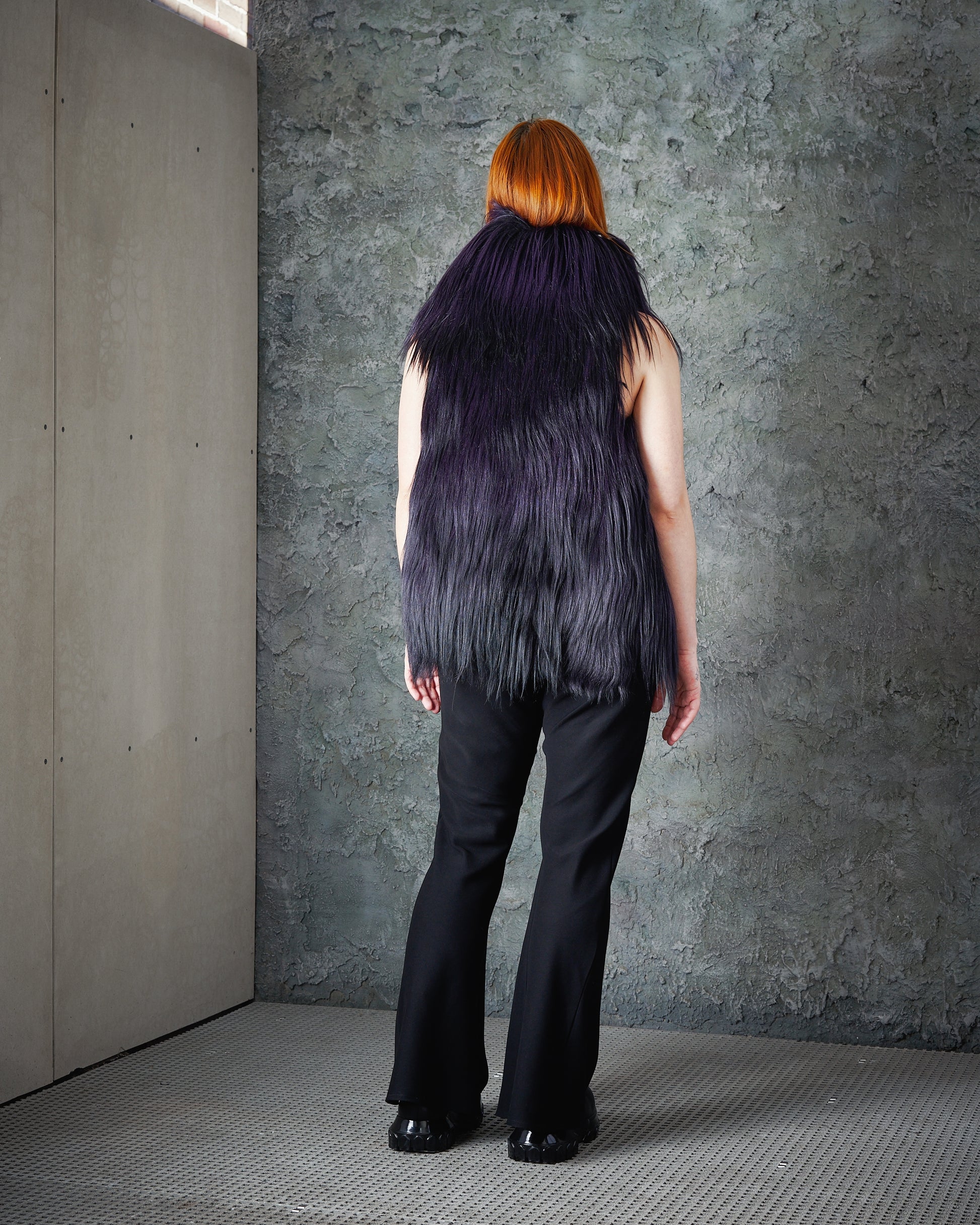 Reverse of a person with red hair wears a voluminous, dark purple Ann Demeulemeester fur vest and black pants against a textured grey wall, exuding a modern, edgy vibe.