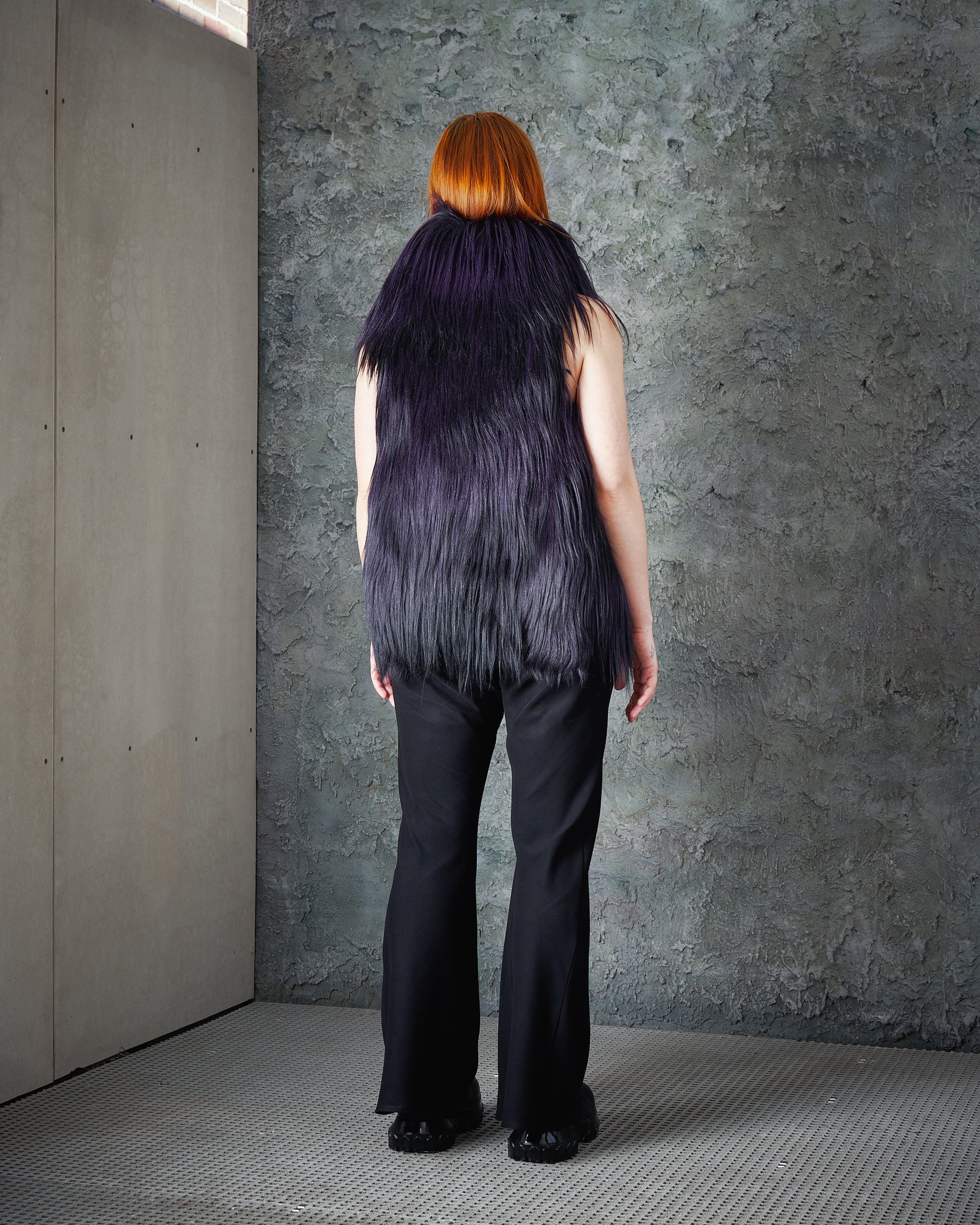 A person with red hair stands facing a textured grey wall, wearing a long, shaggy purple Ann Demeulemeester fur vest and black pants. The scene feels minimalist and modern.