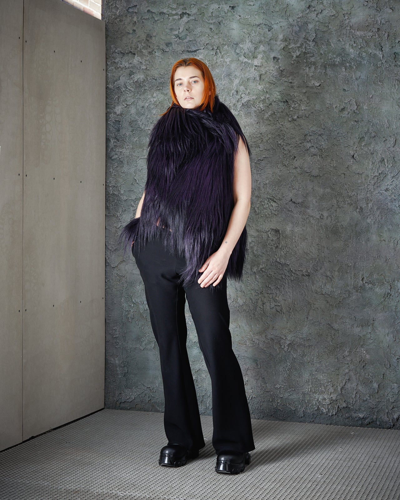 A person with red hair wears a voluminous, dark purple Ann Demeulemeester fur vest and black pants against a textured grey wall, exuding a modern, edgy vibe.