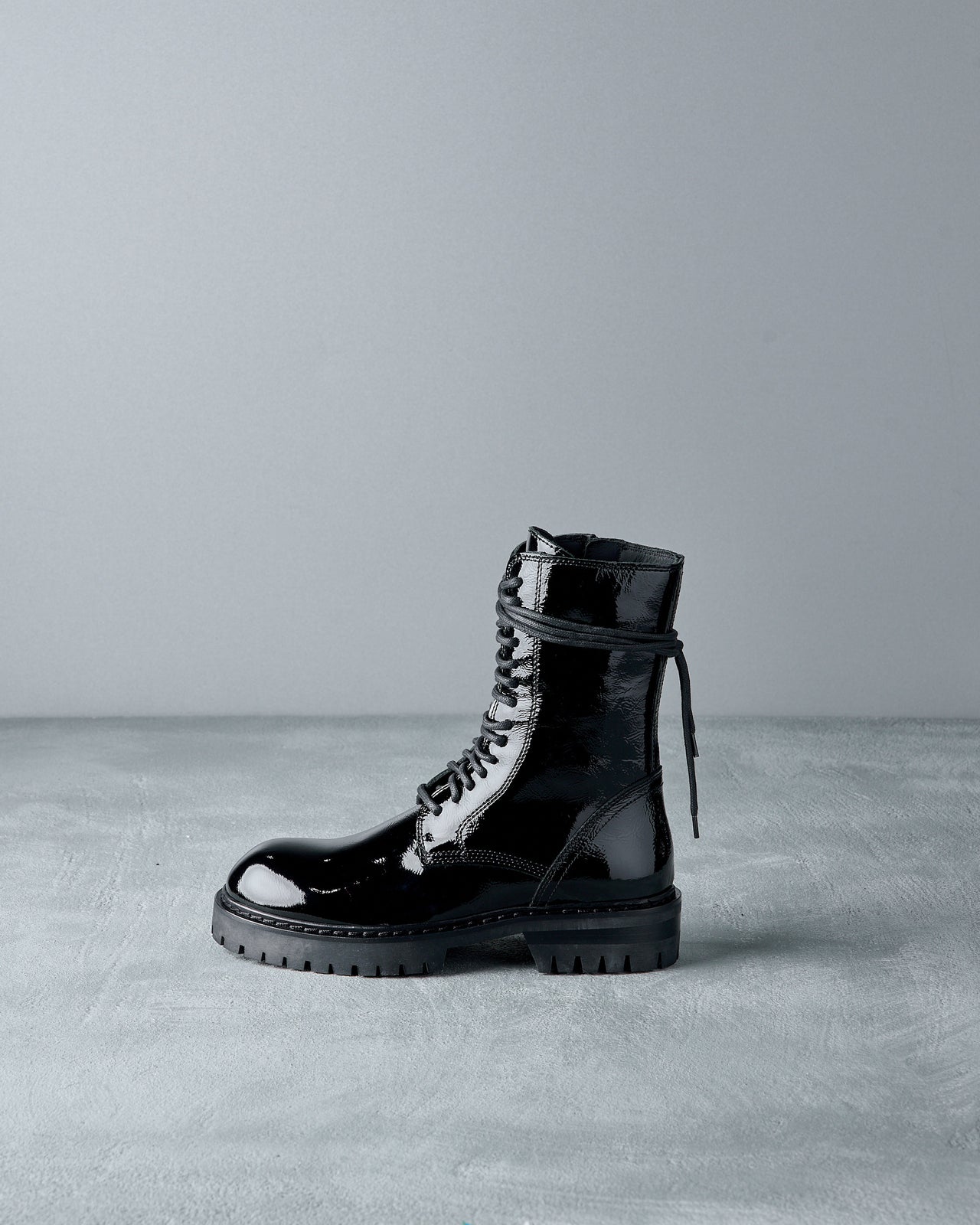 Shiny black Ann Demeulemeester combat boot on a gray surface, featuring lace-up design and rugged sole. The lighting highlights the boot’s glossy finish.