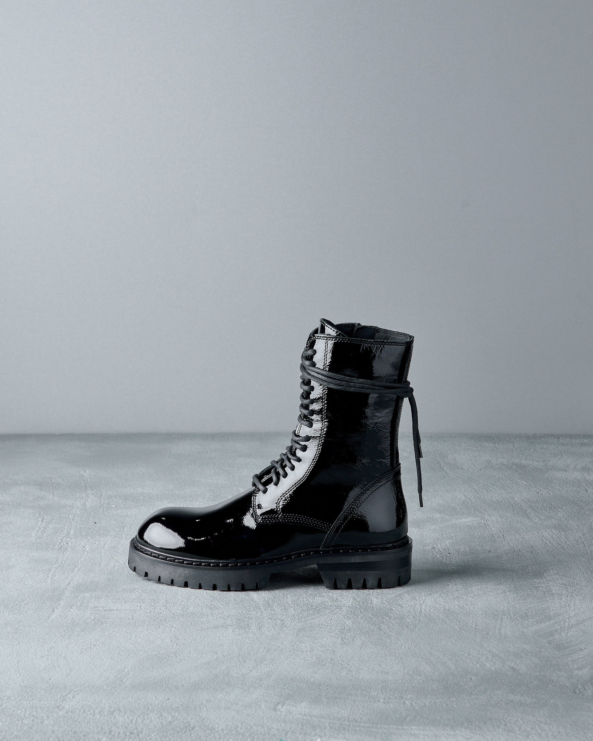 Shiny black Ann Demeulemeester combat boot on a gray surface, featuring lace-up design and rugged sole. The lighting highlights the boot’s glossy finish.
