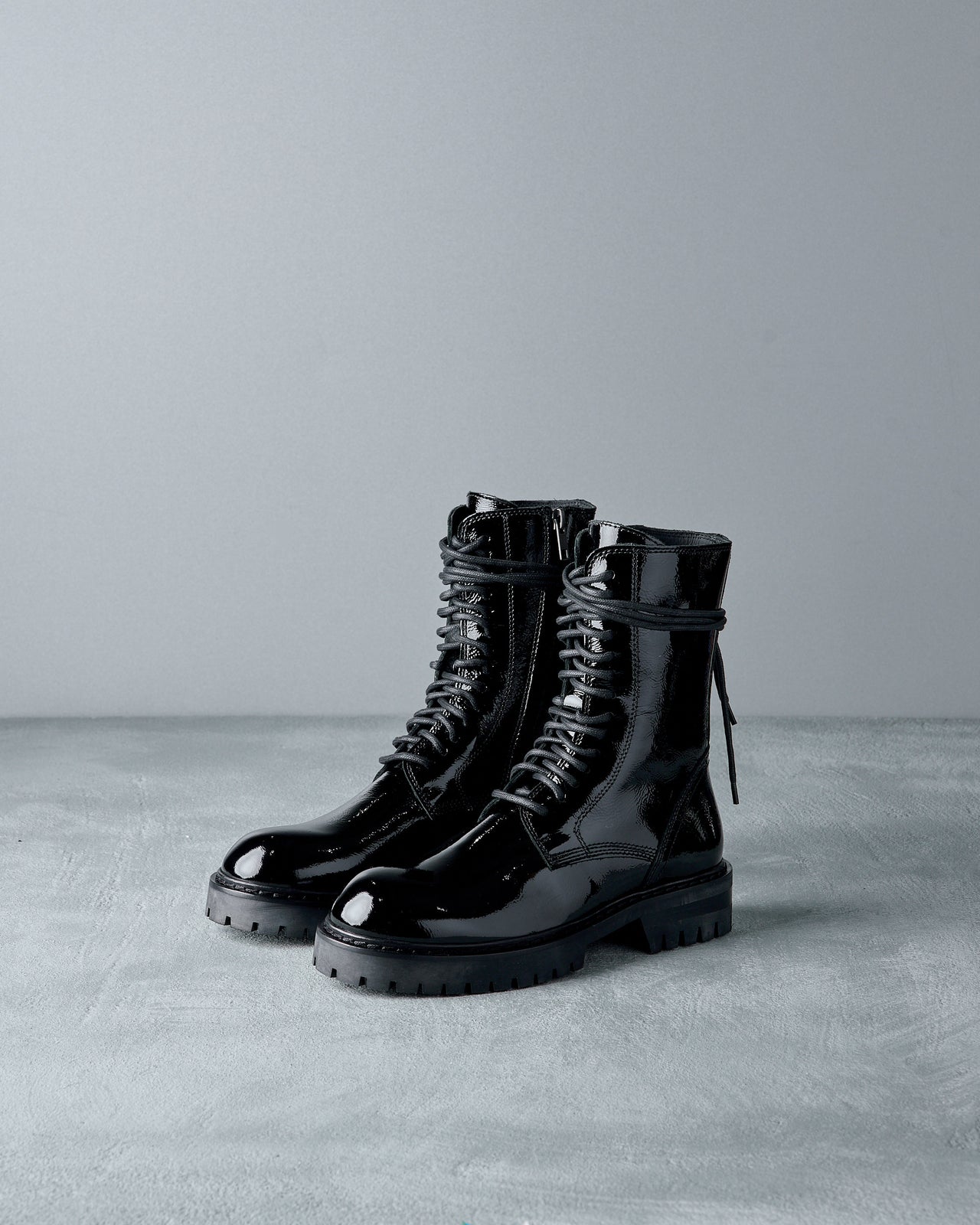 Shiny black Ann Demeulemeester combat boot on a gray surface, featuring lace-up design and rugged sole. The lighting highlights the boot’s glossy finish.