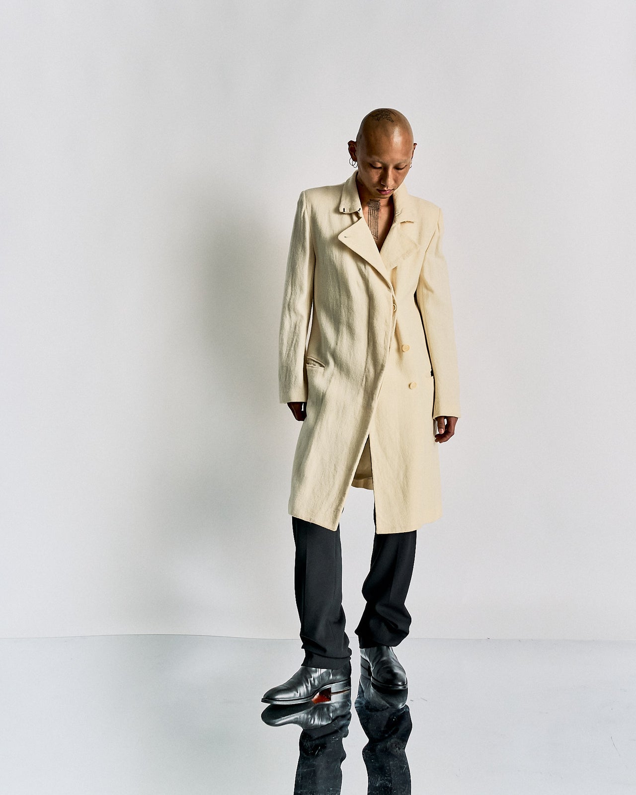 A person standing in front of a white background, wearing a beige double-breasted Ann Demeulemeester wool coat with hidden button closures, two external pockets, and single-button cuffs.