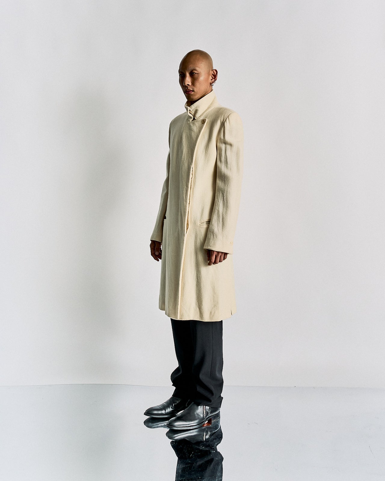 A person standing in front of a white background, wearing a beige double-breasted Ann Demeulemeester wool coat with hidden button closures, two external pockets, and single-button cuffs.